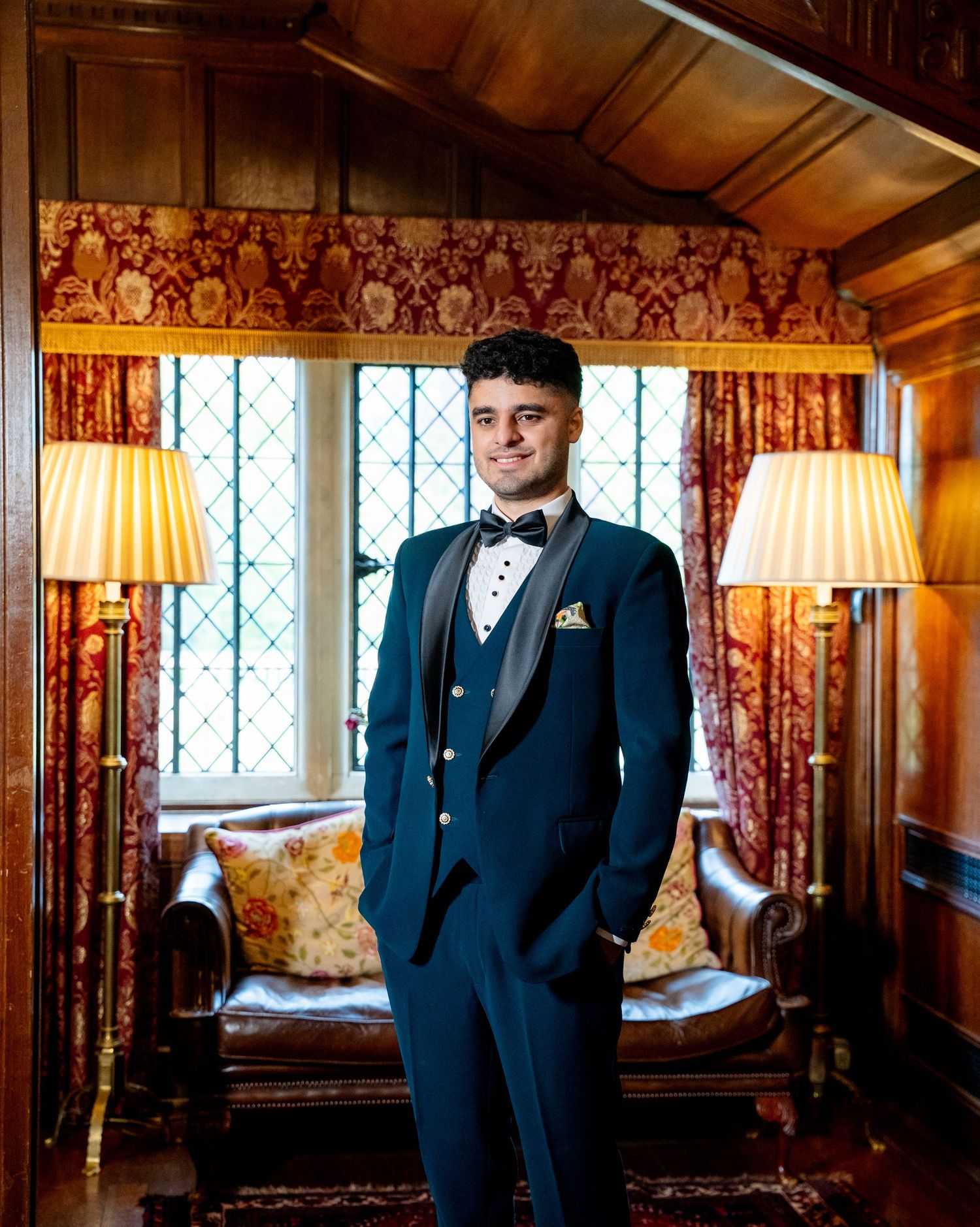 Young man in navy tuxedo with bow tie standing in ornate wood-paneled room with vintage decor.
