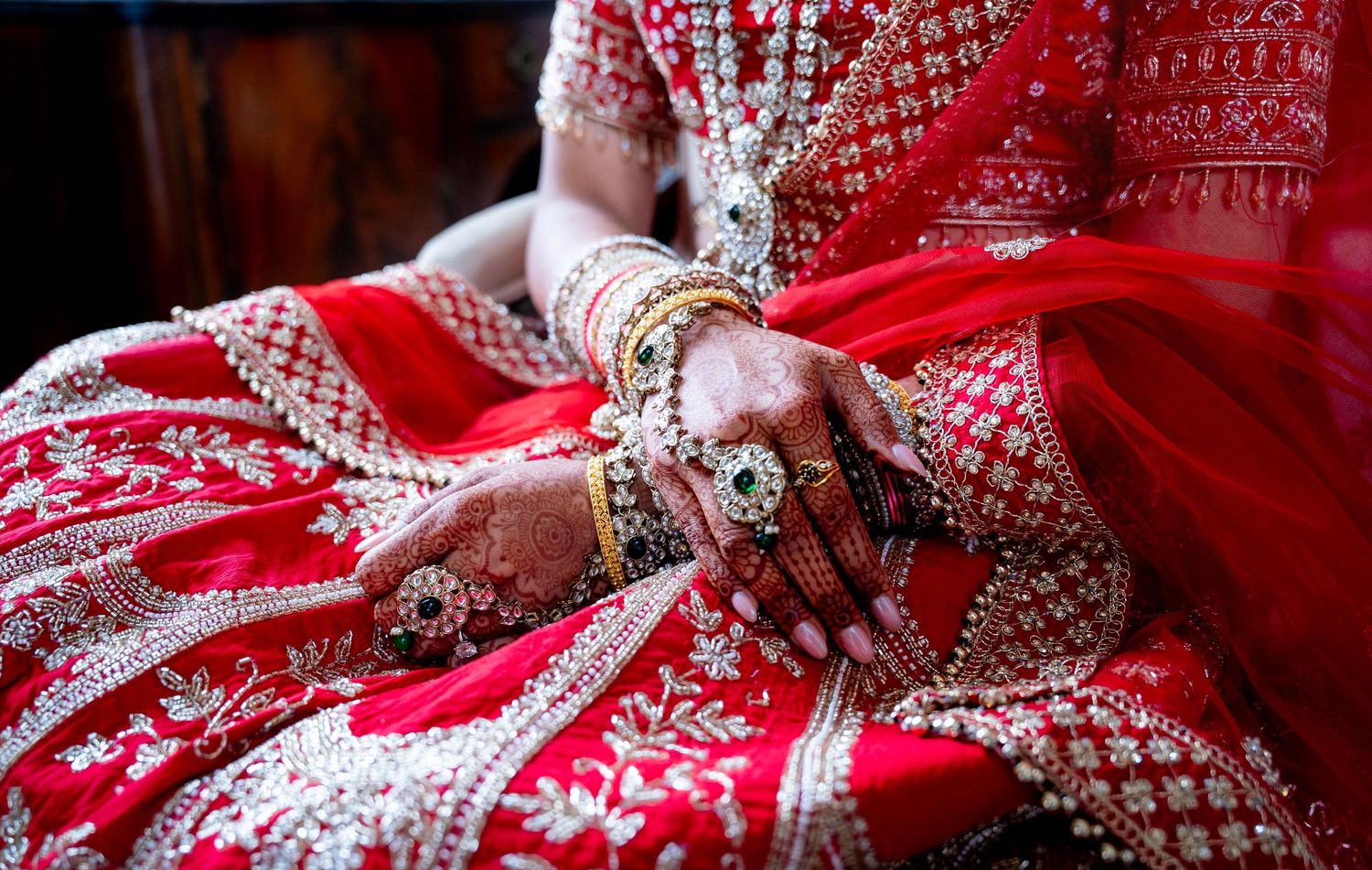 Close-up of Indian bride's hands adorned with mehndi, gold bangles, and jeweled bracelets on red lehenga.