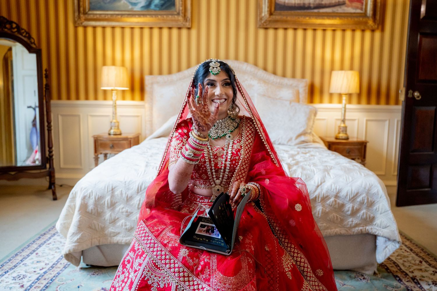 Indian bride in red lehenga and jewelry smiles while holding tablet on bed in elegant room.
