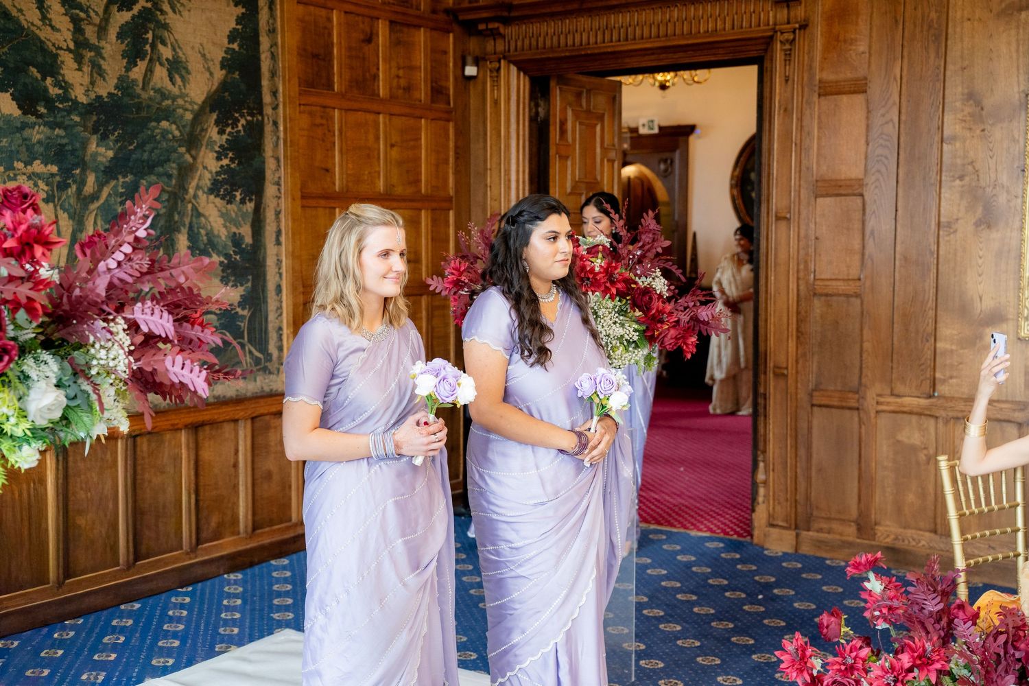 Two bridesmaids in lavender dresses holding bouquets stand in an elegant wood-paneled hall with red floral arrangements.