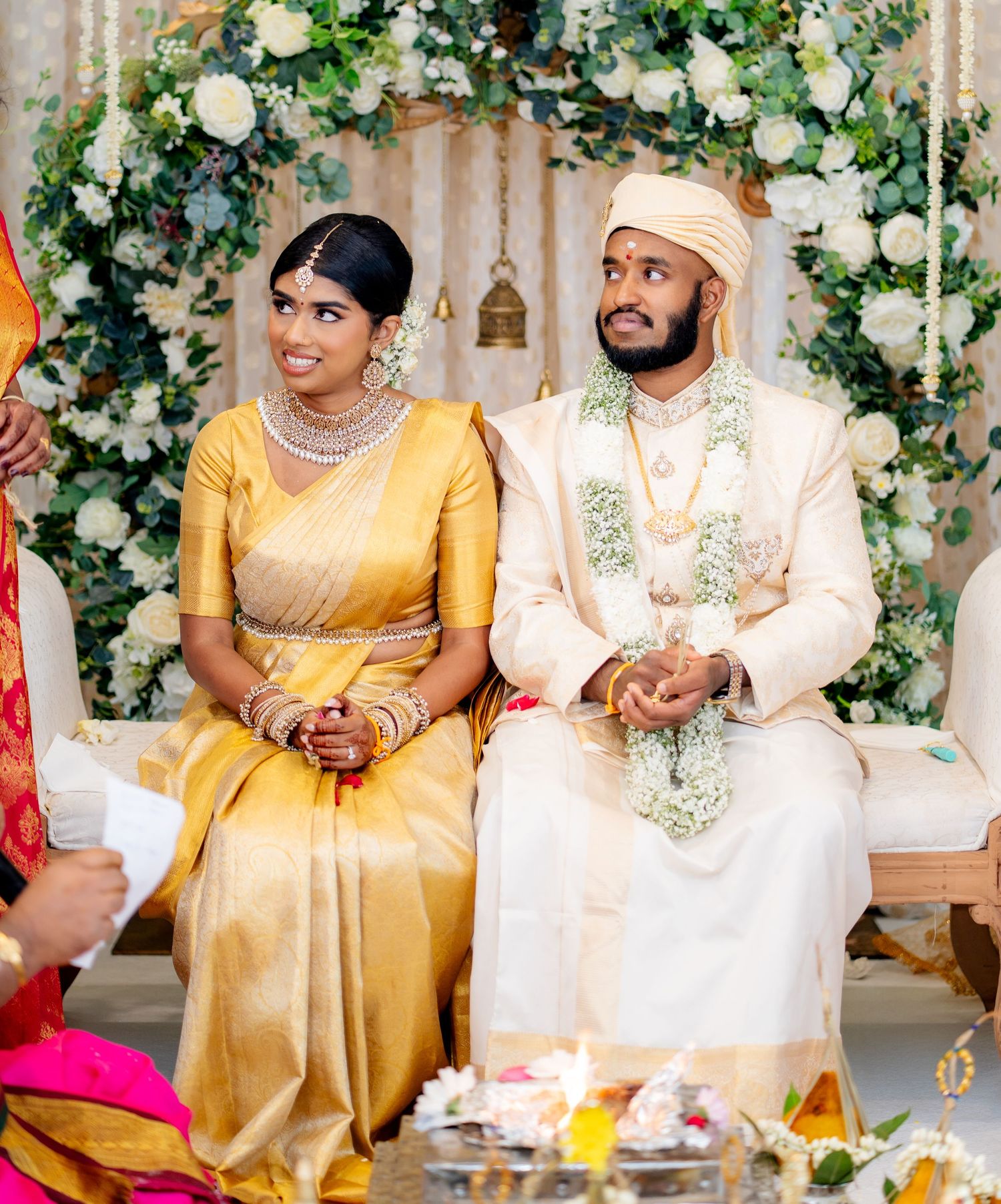 South Indian bride in gold Kanjivaram saree and groom in cream sherwani seated together at traditional wedding mandap.