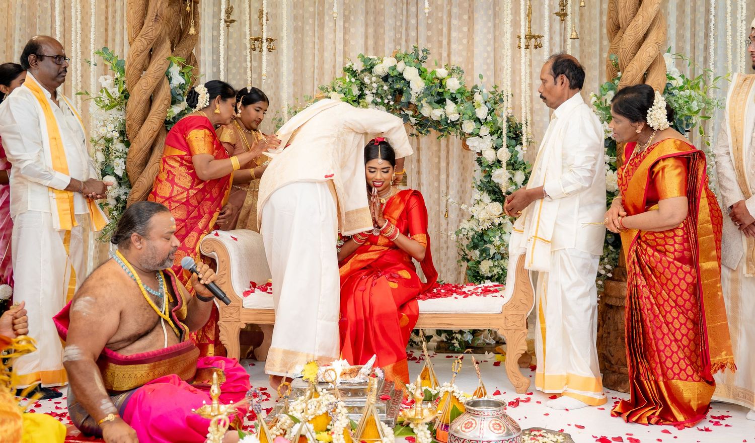 Bride in red saree sits during traditional South Indian Hindu wedding ceremony with priest and family gathered around.