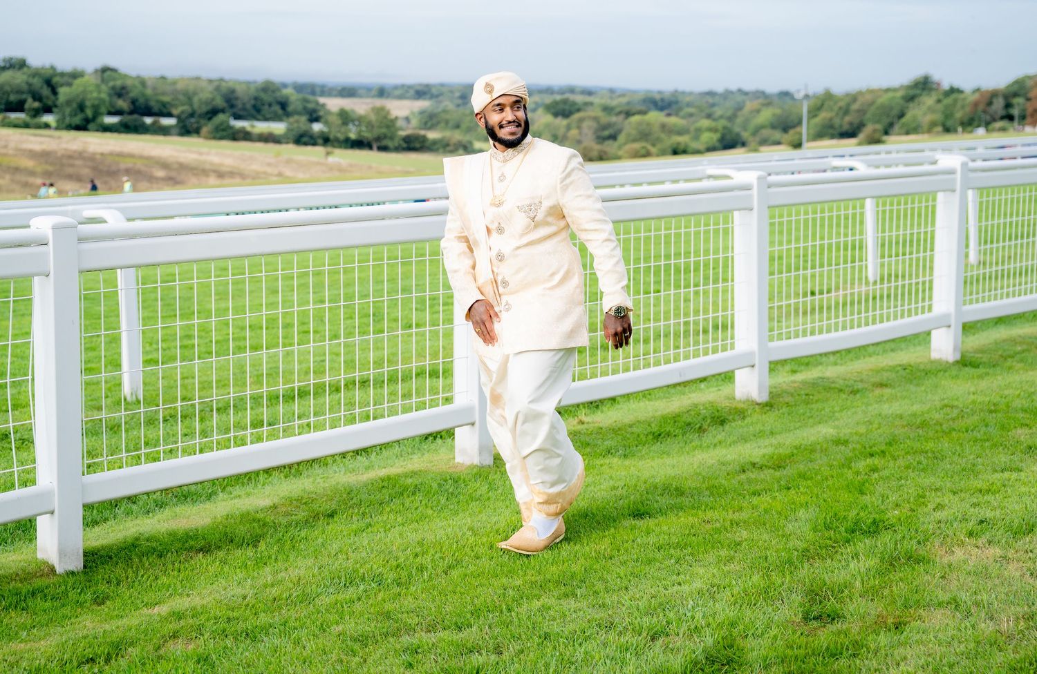 Man in traditional South Asian sherwani and turban walking on green grass by white fence at racecourse.