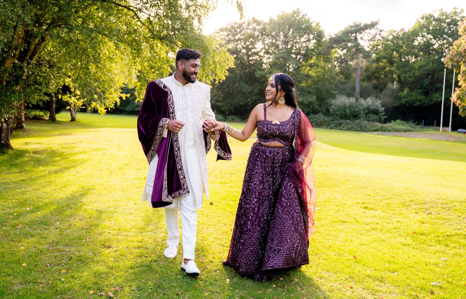 Smiling Indian wedding couple in purple and white traditional outfits walking on green lawn at golden hour.
