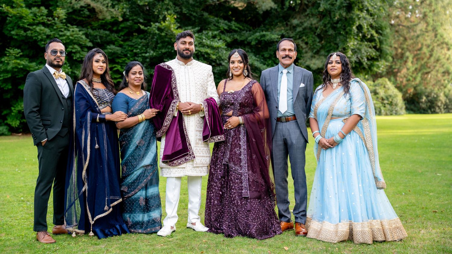 Indian wedding party of seven posing outdoors in traditional attire including purple lehenga and blue sarees on green lawn.