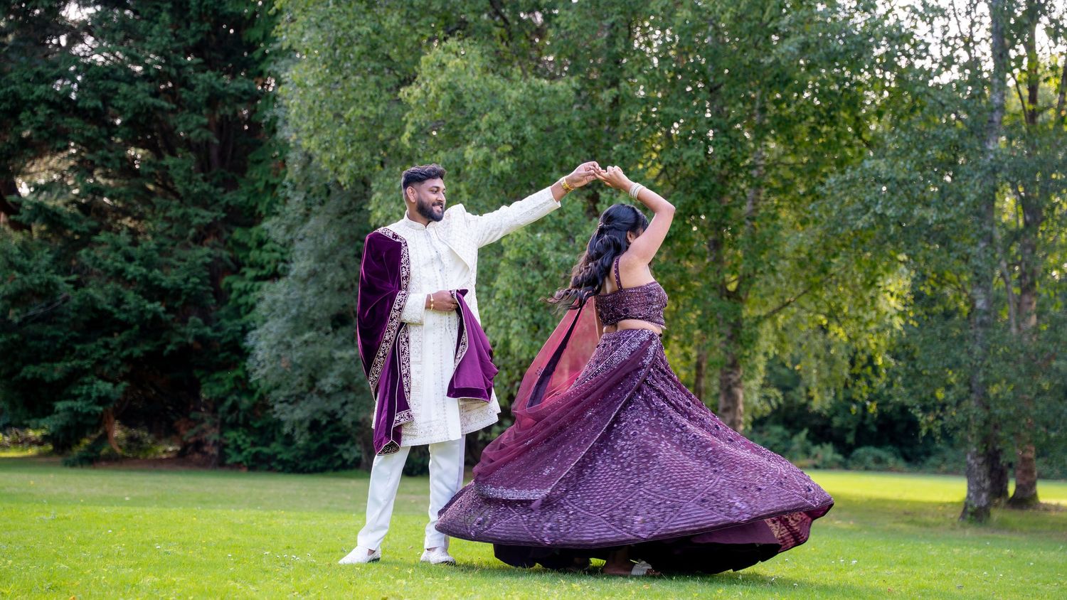 Indian bride in purple lehenga twirling while groom in white sherwani holds her hand outdoors.