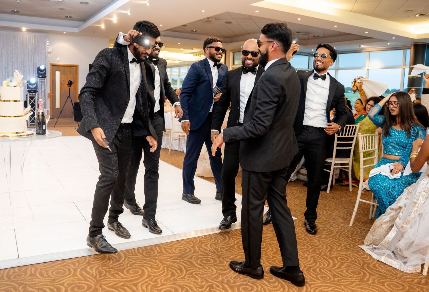 Groomsmen in black tuxedos dancing together on the dance floor at an elegant wedding reception.