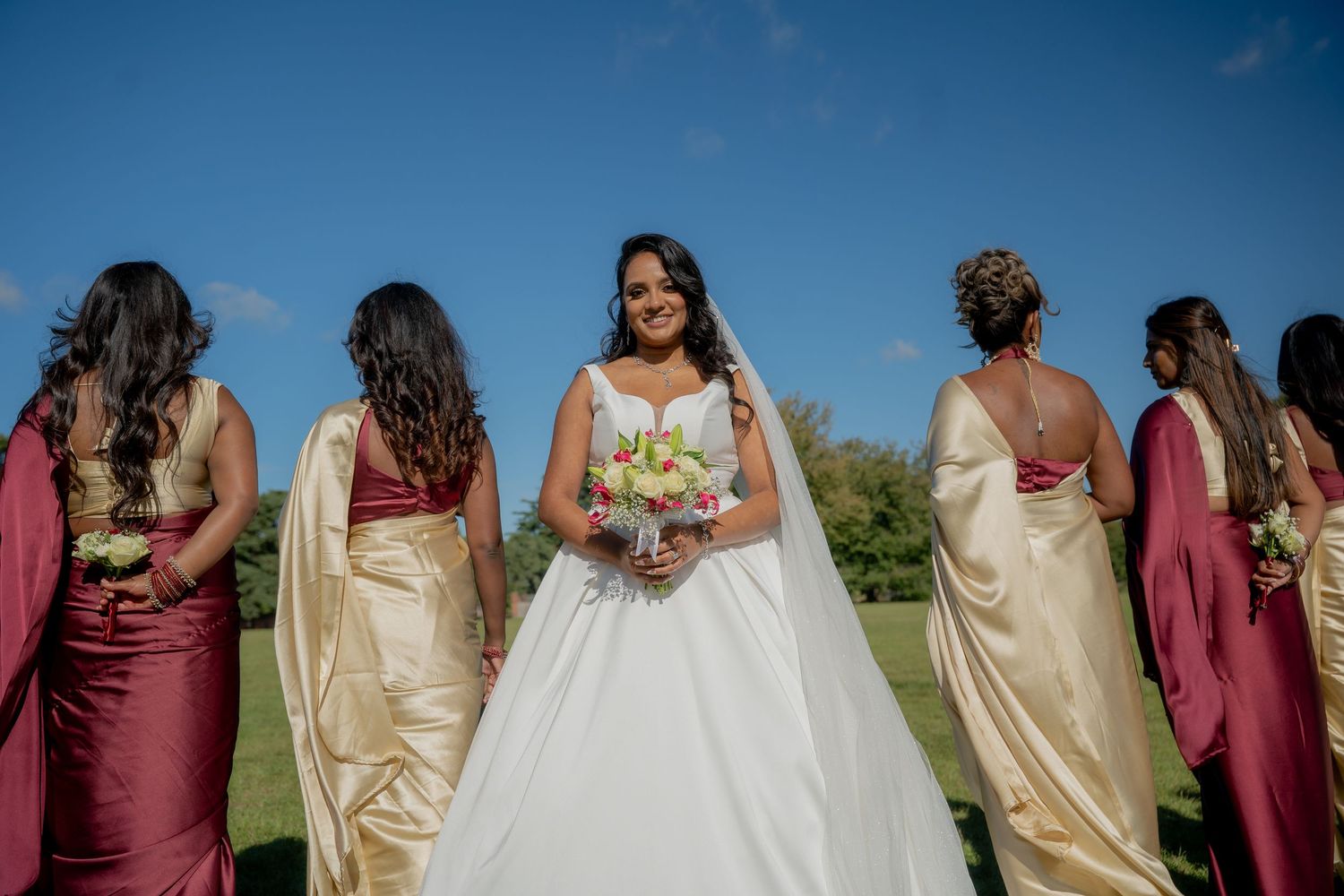 Smiling bride in white gown holds bouquet while bridesmaids in gold and burgundy dresses stand behind her.