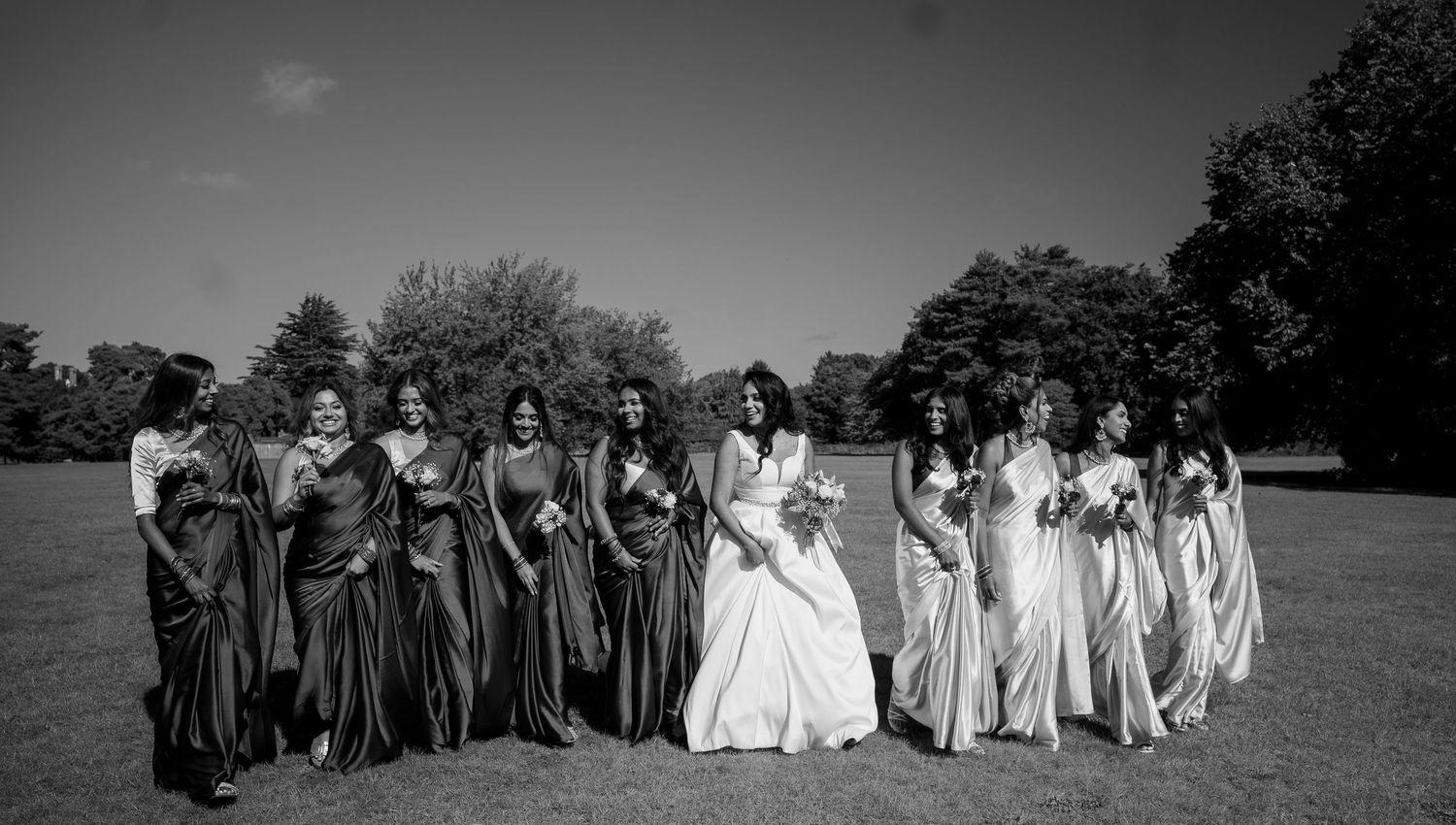 Black and white photo of a bride with her bridesmaids in long gowns posing outdoors on a grassy field.