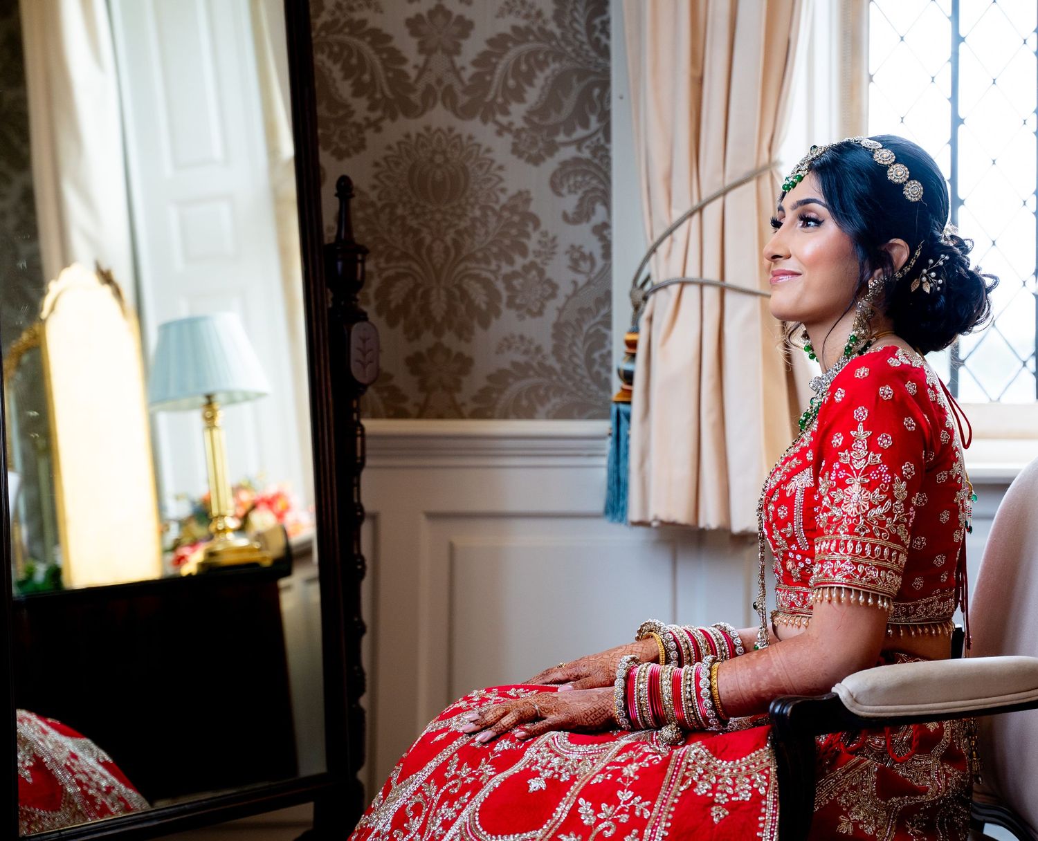 Beautiful Indian bride in red bridal lehenga seated beside ornate mirror in elegant dressing room.