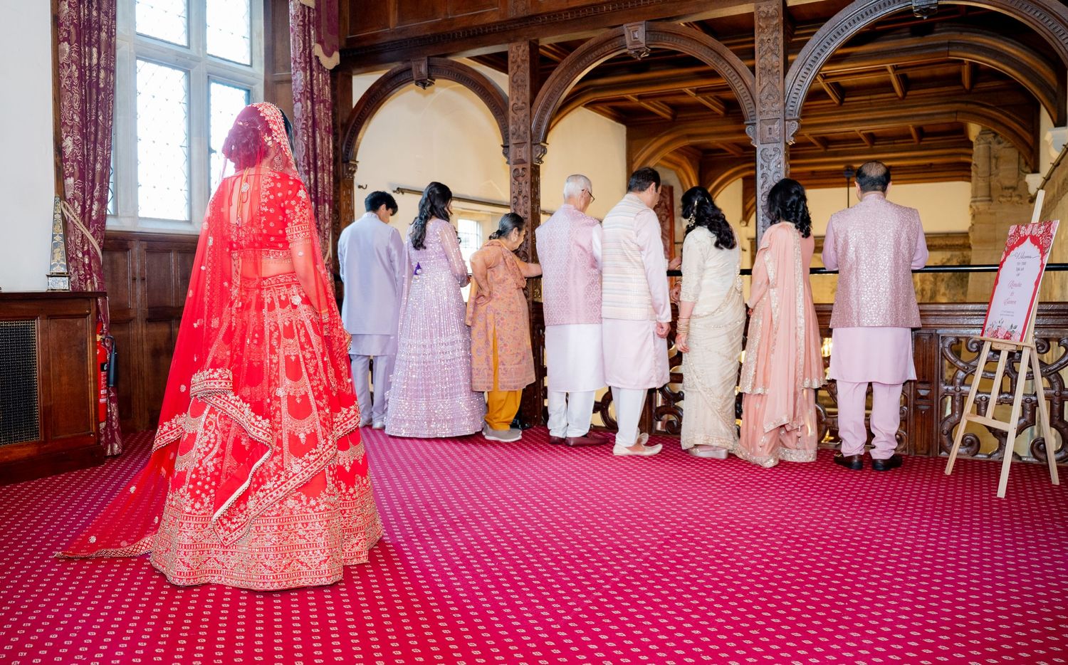 Bride in red lehenga walks toward family group in ornate hall during Indian wedding ceremony.