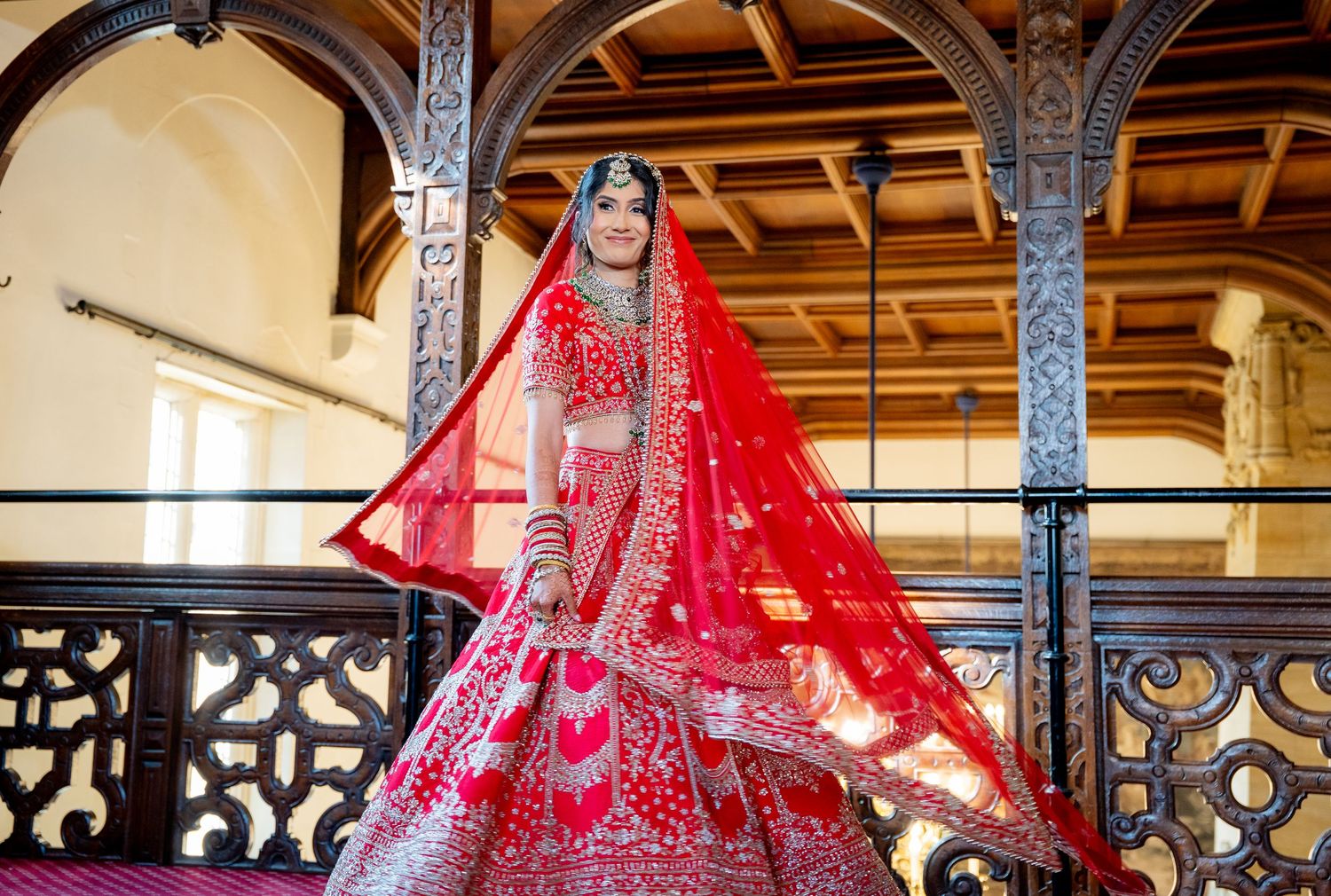 Indian bride in red lehenga posing on balcony of traditional wooden venue.