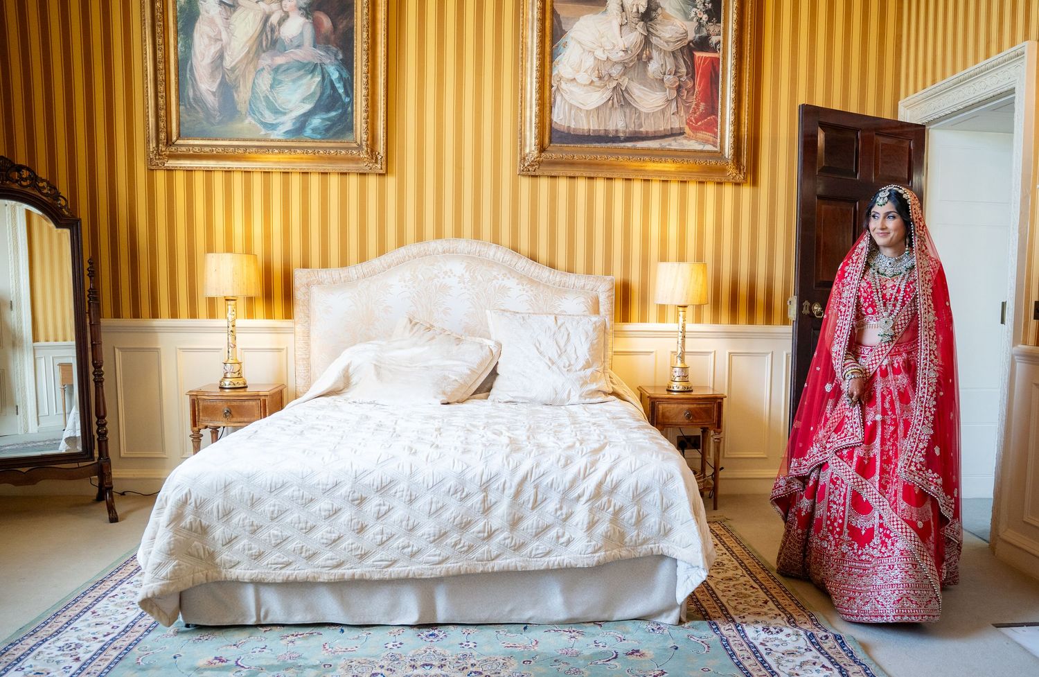 Indian bride in red lehenga standing beside elegant white bed in gold-wallpapered bridal suite.