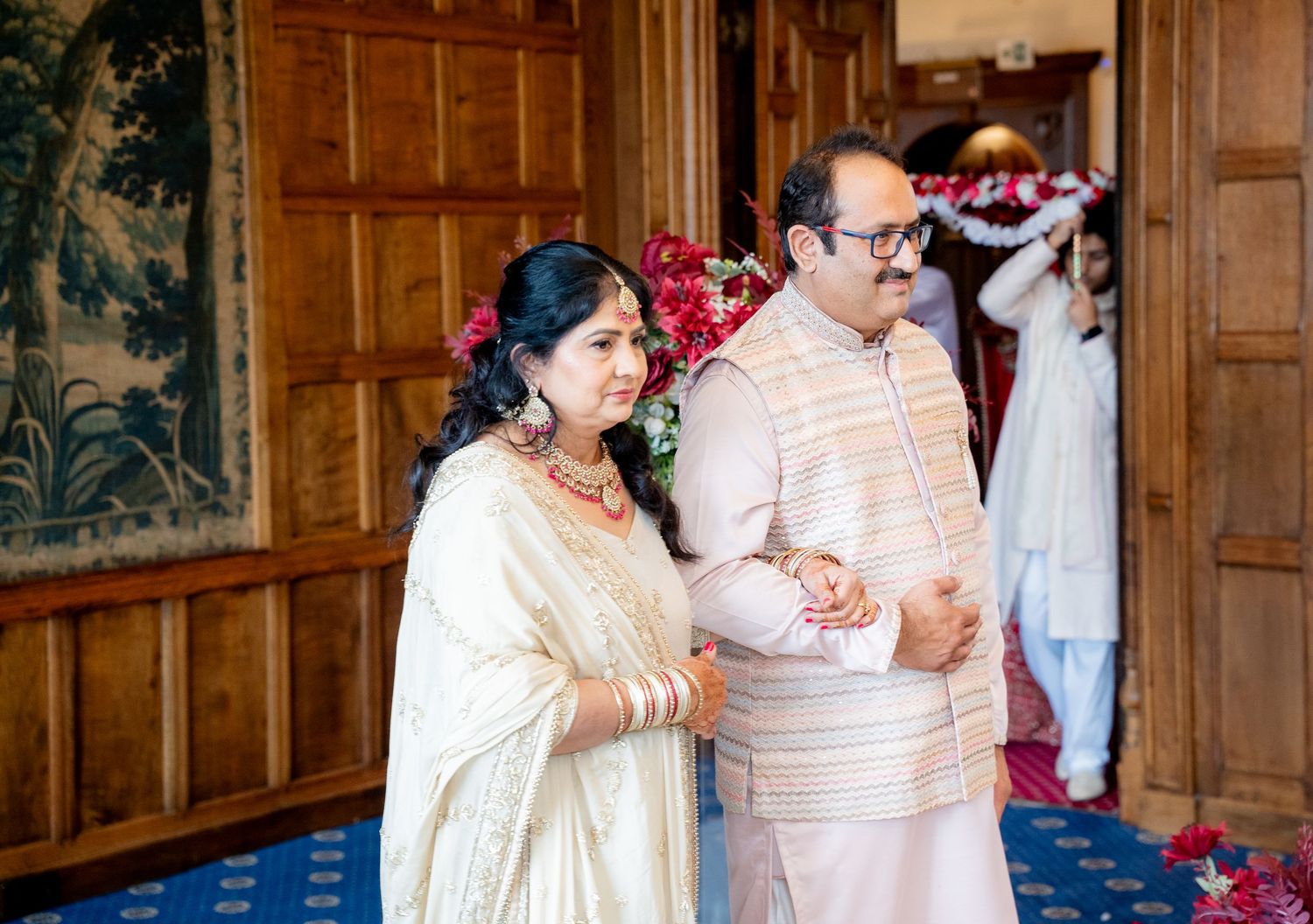 Couple in traditional Indian wedding attire walk arm in arm through decorated venue corridor.