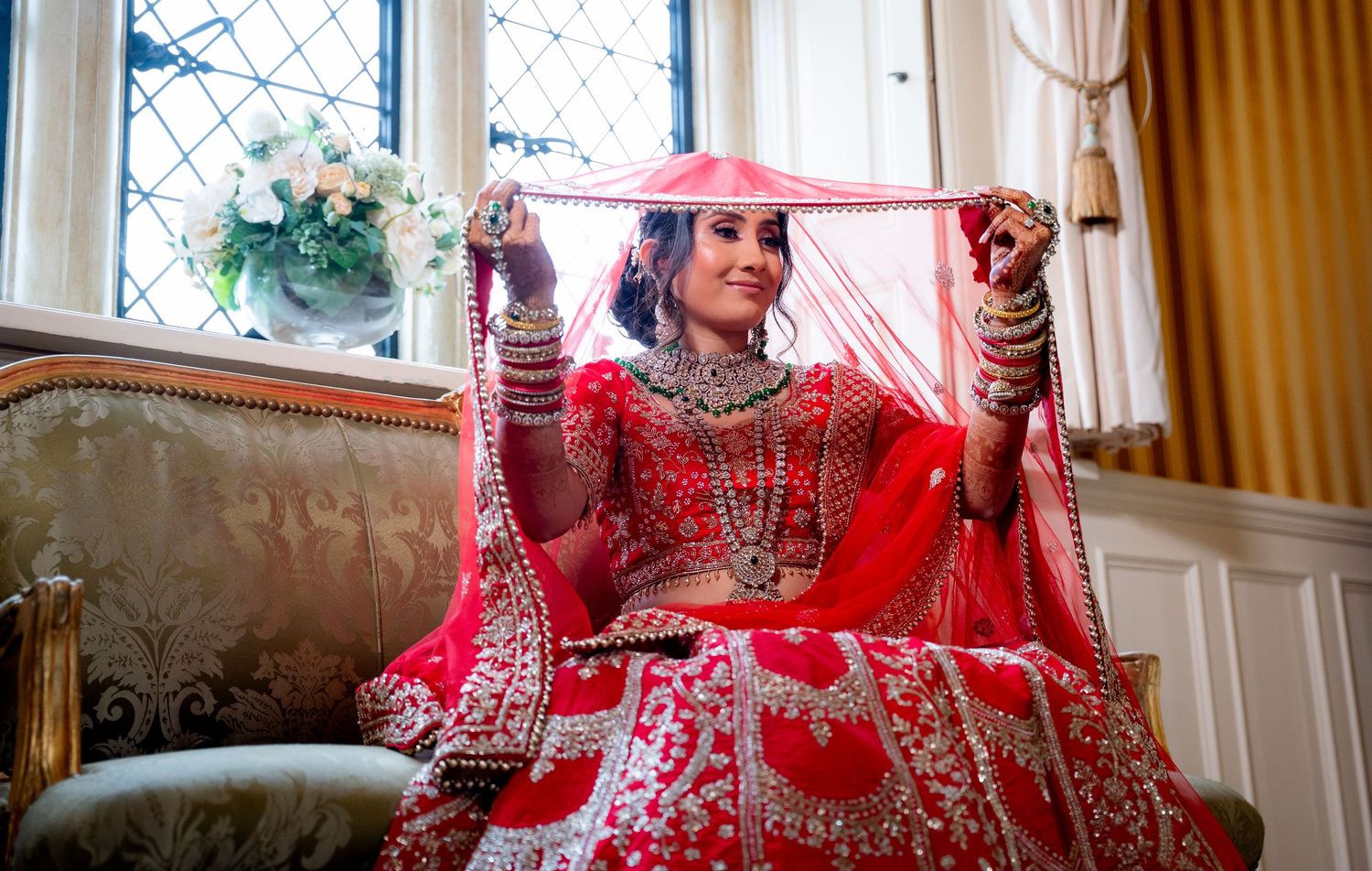 Indian bride in red lehenga holds embroidered dupatta veil wide open near ornate window.