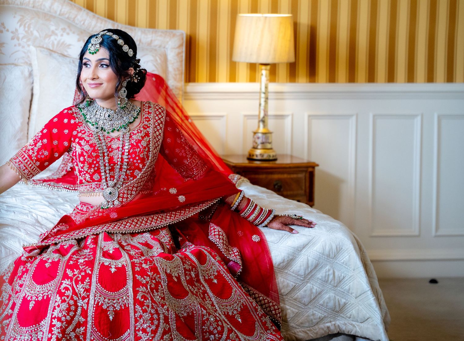 Indian bride in ornate red lehenga with heavy embroidery sits gracefully on white bed.