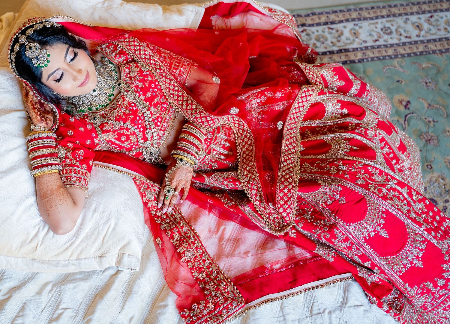 A bride in an ornate red lehenga with gold embroidery lies gracefully, adorned with traditional jewelry and bridal makeup.