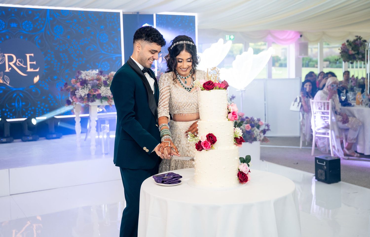 Bride and groom in traditional South Asian wedding attire cutting white floral wedding cake at reception.