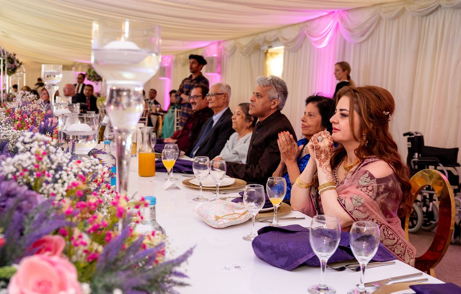 Elegant wedding reception dinner with guests seated at a floral-decorated table inside a white marquee tent.