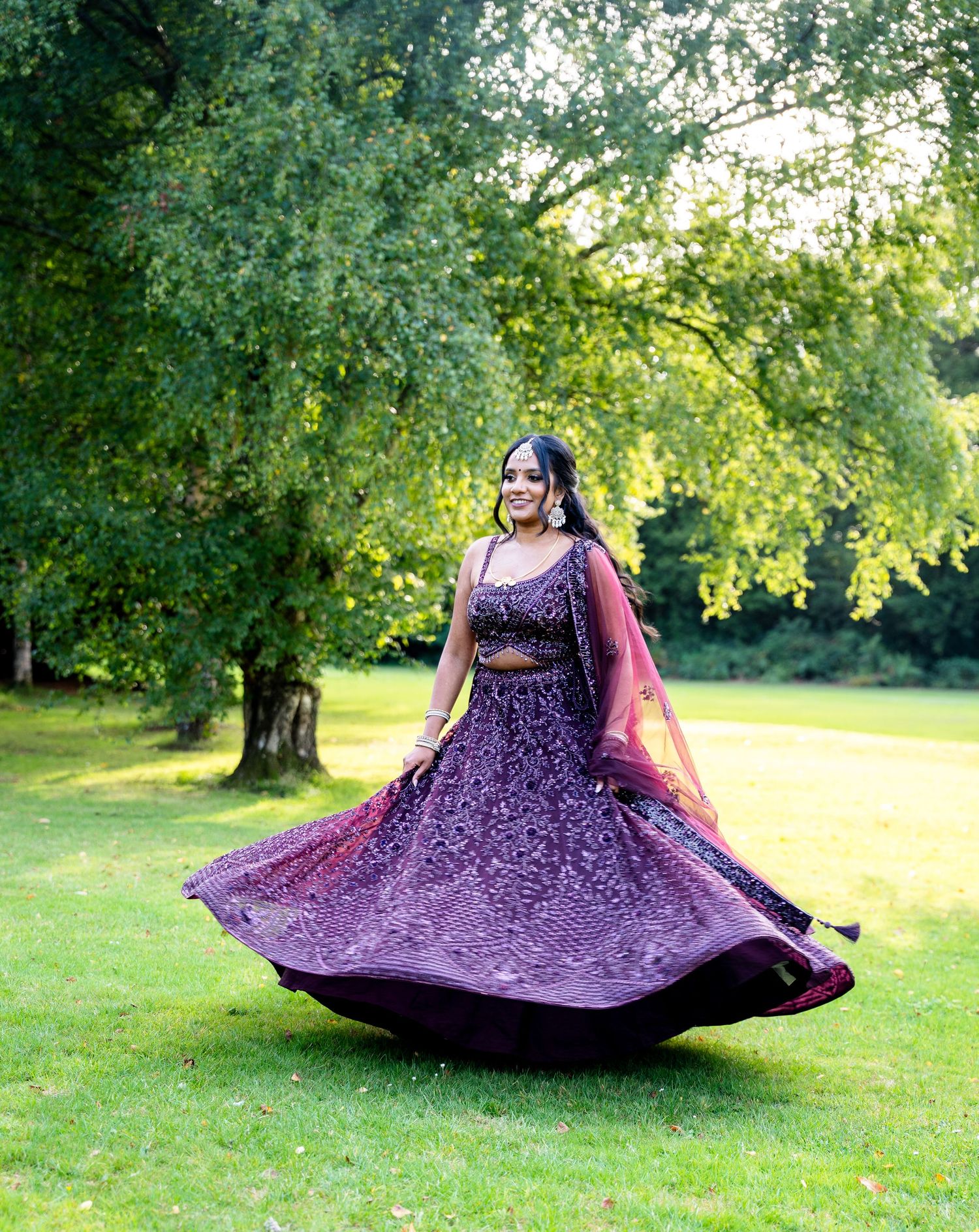 A woman twirls in a stunning purple embroidered lehenga outdoors, smiling joyfully in a lush green park setting.