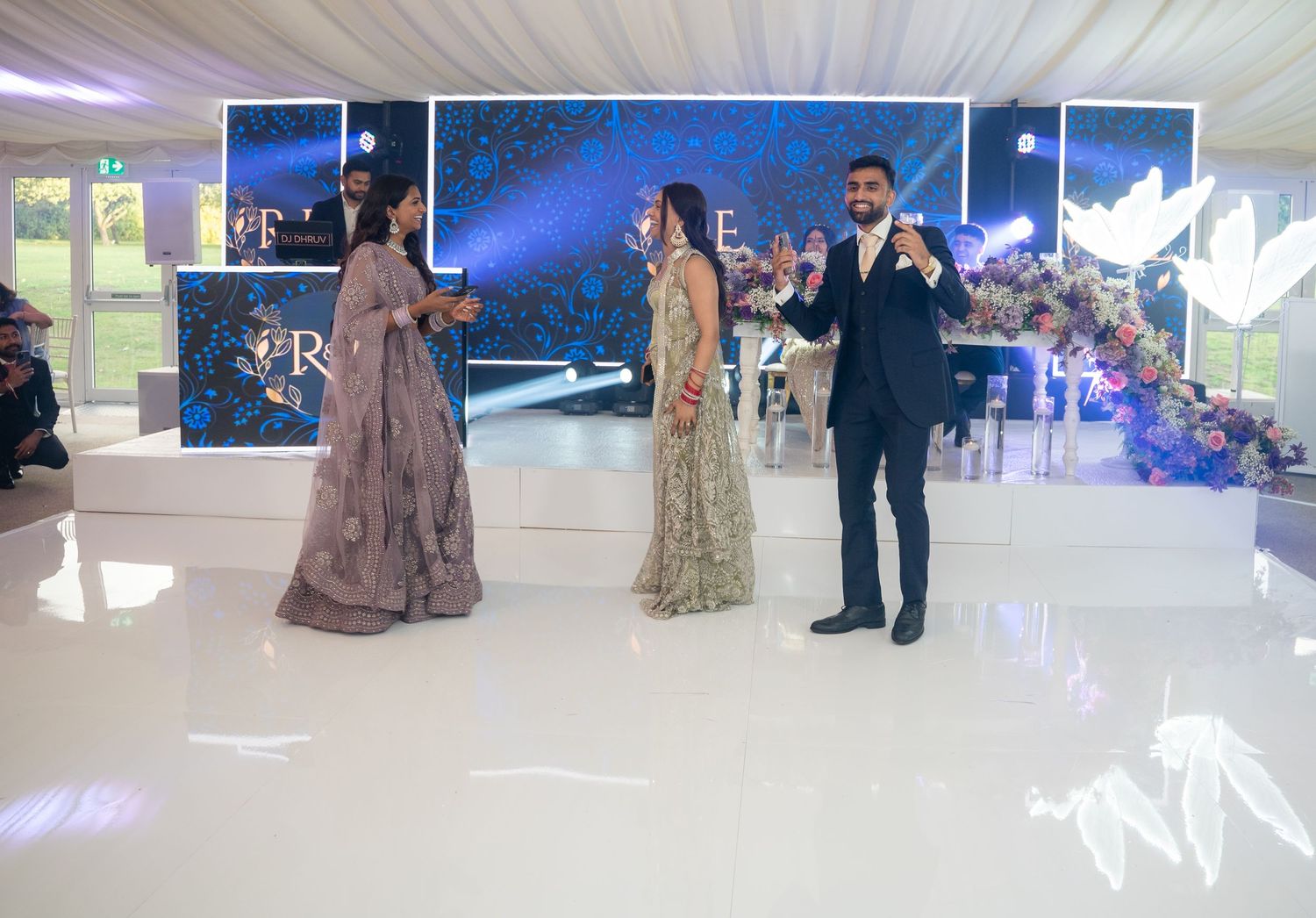 Elegant wedding reception on a white dance floor with guests in formal attire, floral backdrop, and soft blue lighting.