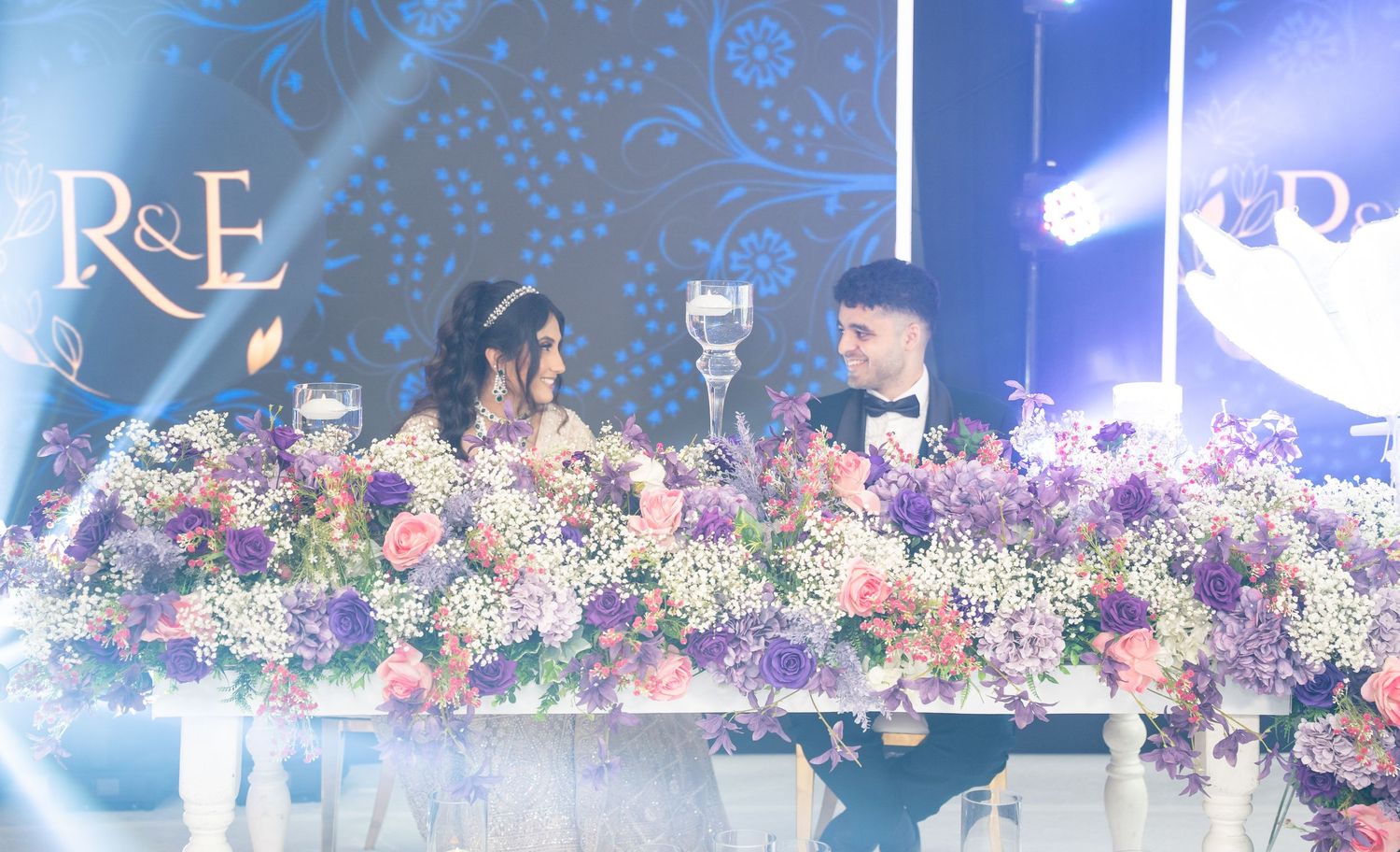 Bride and groom seated at floral wedding reception table with purple and pink flowers under elegant blue lighting.