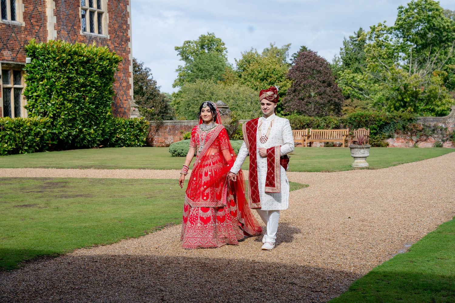 Indian bride and groom in red and white traditional outfits walk through manicured gardens of a historic estate.