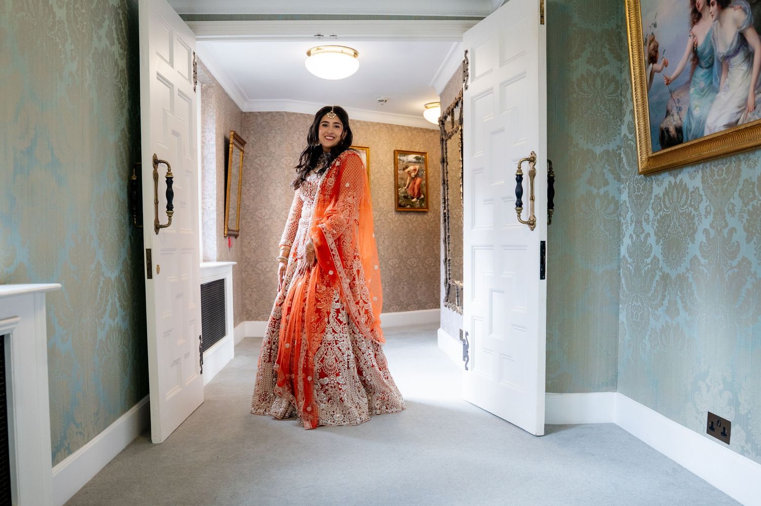 Indian bride in vibrant orange lehenga stands in doorway of elegant wallpapered hallway.
