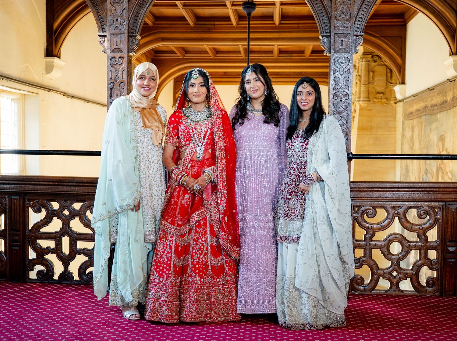 Four women in elegant South Asian attire pose together in an ornate venue, with a bride in red lehenga at center.