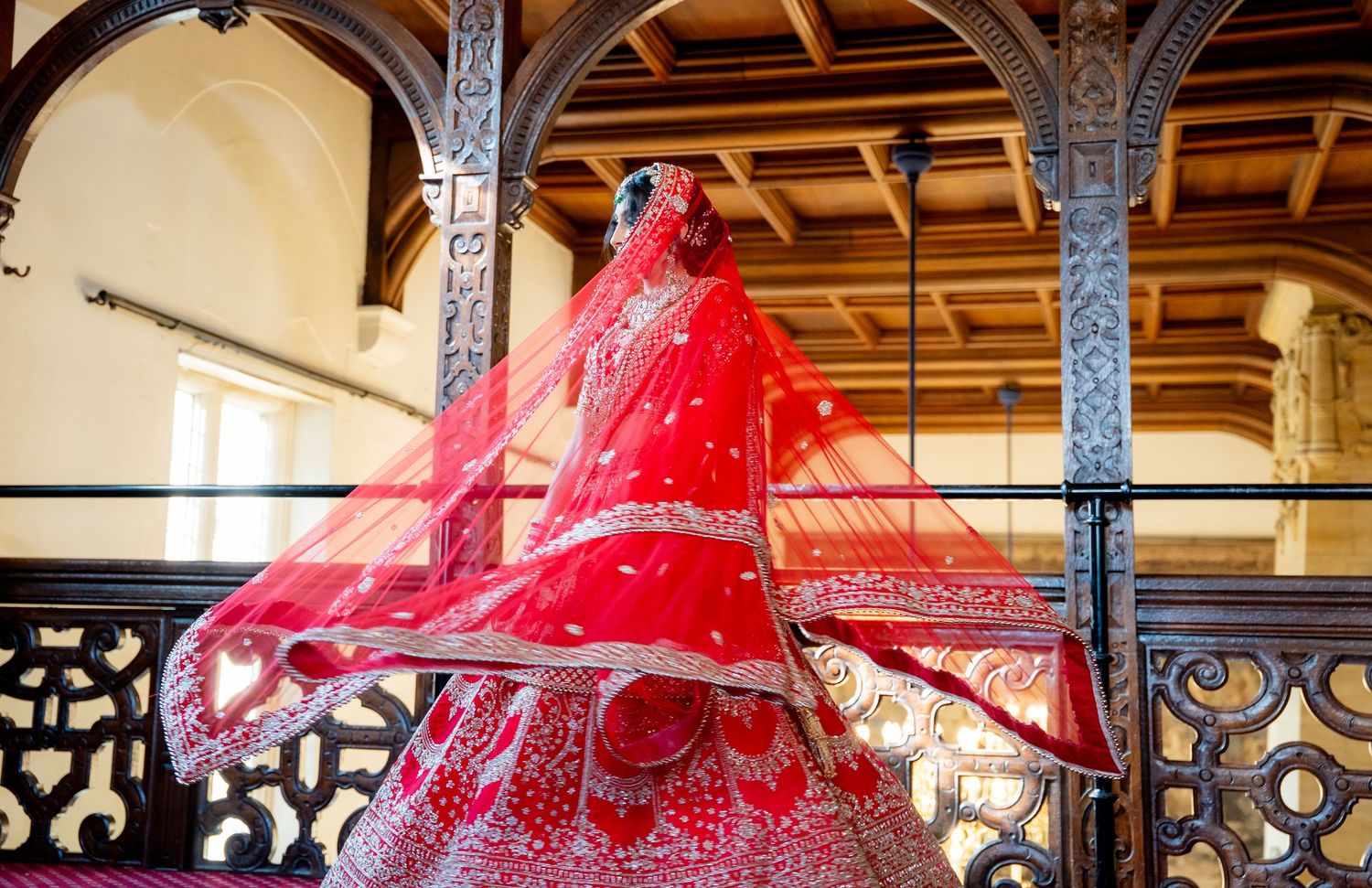Indian bride in red embroidered lehenga with flowing dupatta captured from below on carved balcony.
