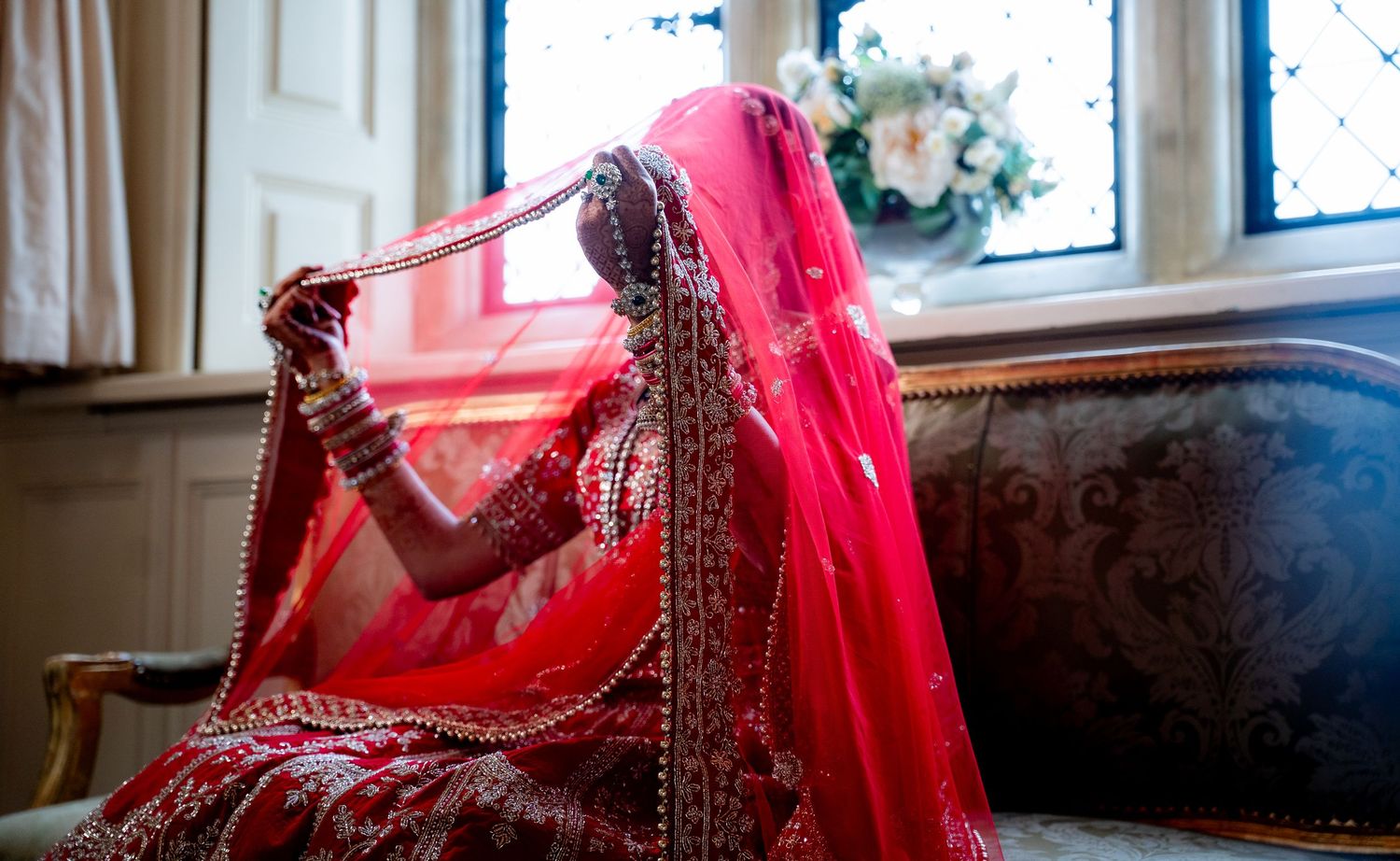 Indian bride in red bridal lehenga adjusting dupatta veil while seated on ornate sofa.
