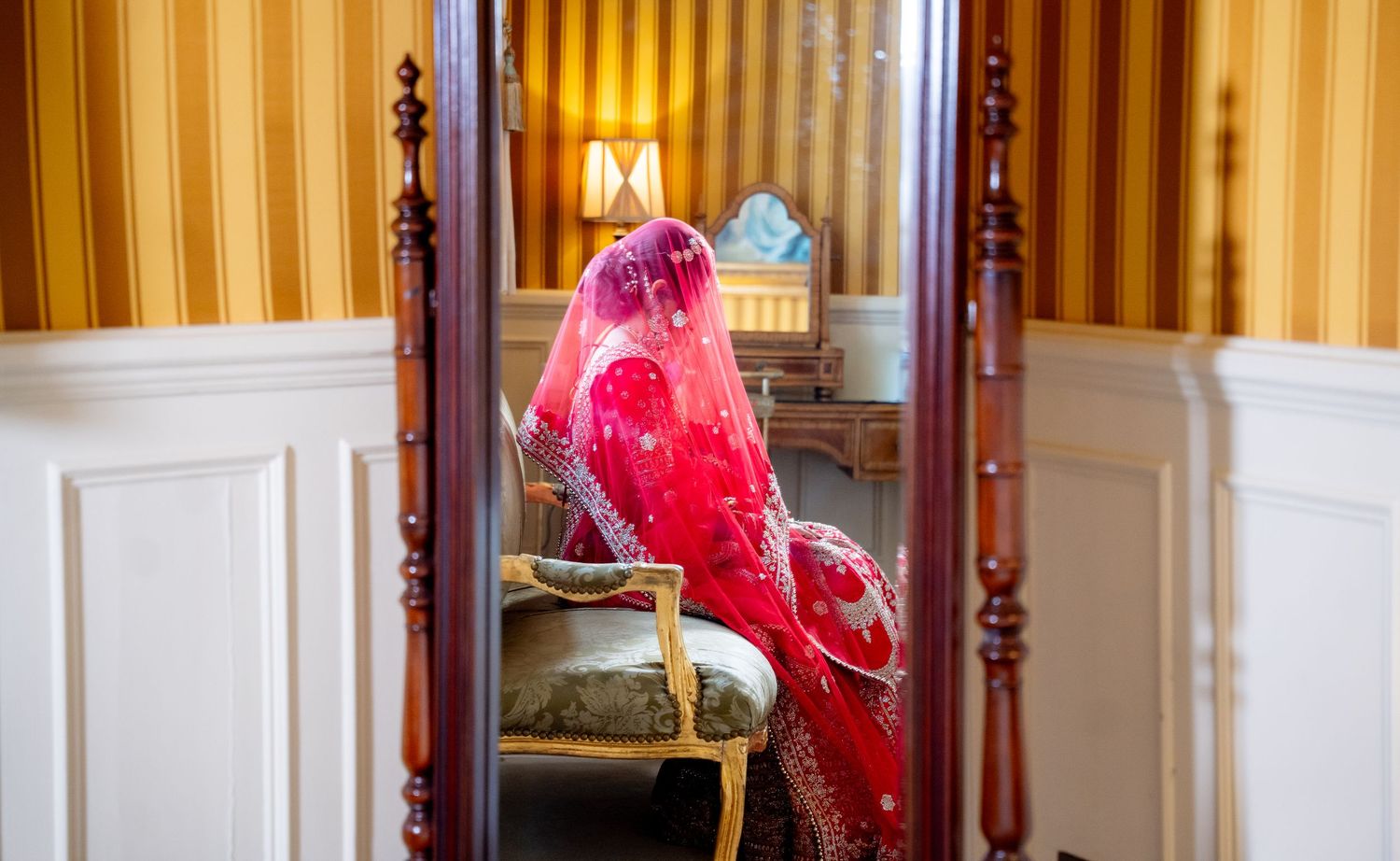 Indian bride in vibrant red lehenga reflected in ornate mirror during bridal preparation.