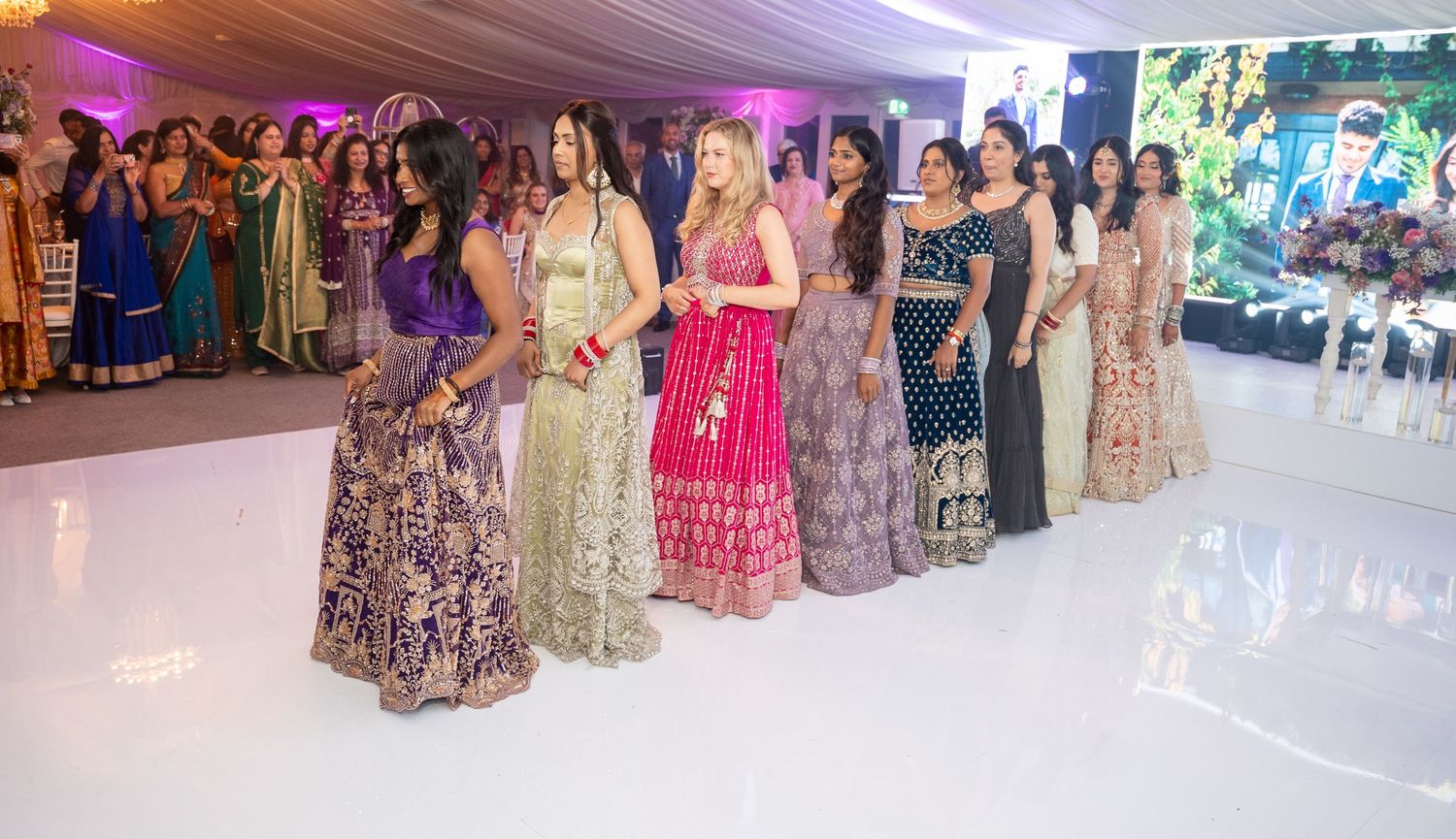 Women in colorful Indian lehengas line up on a white dance floor at a vibrant Indian wedding reception.