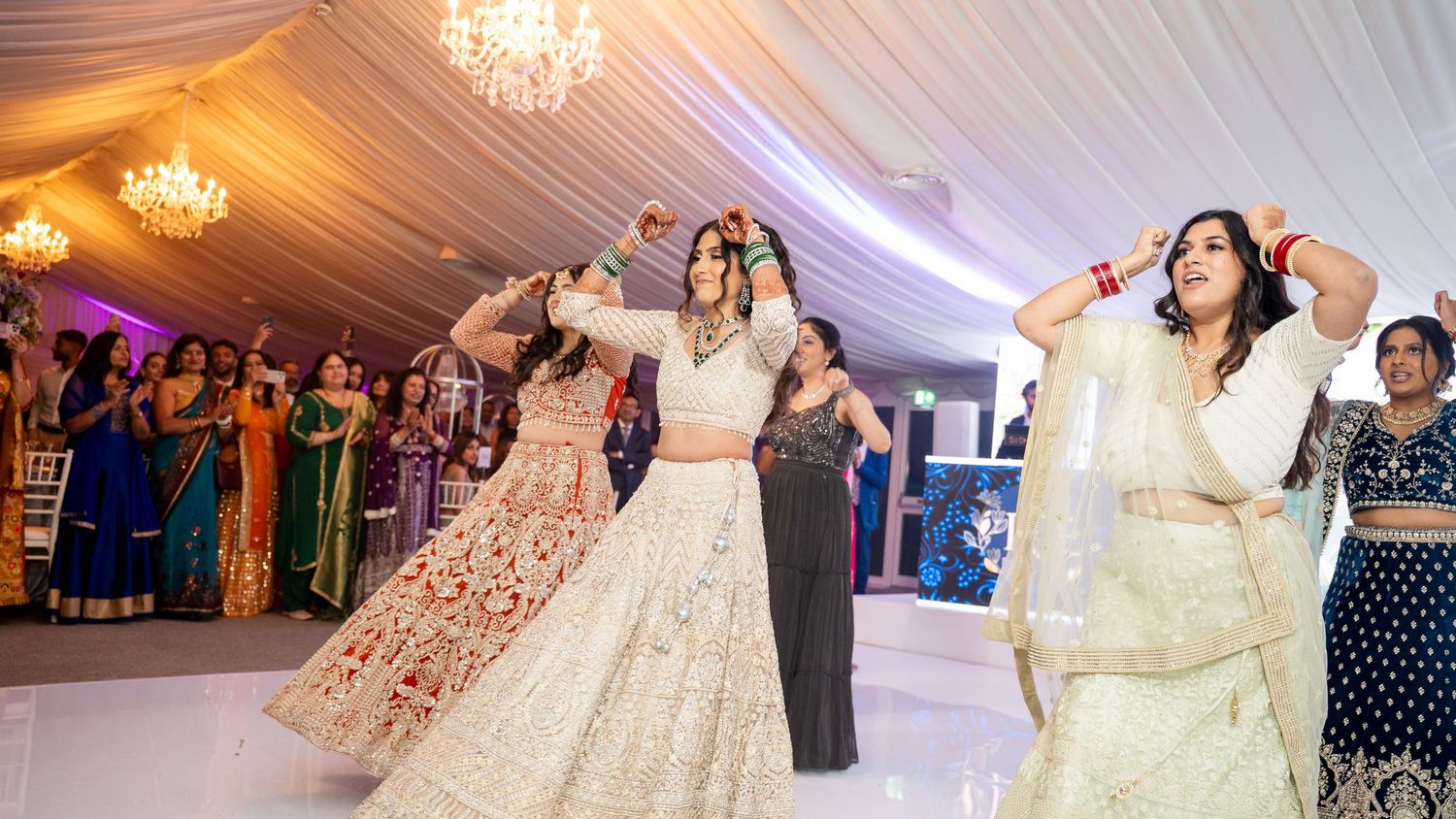 Women in colorful traditional Indian attire perform a dance at a festive wedding celebration inside a decorated tent venue.