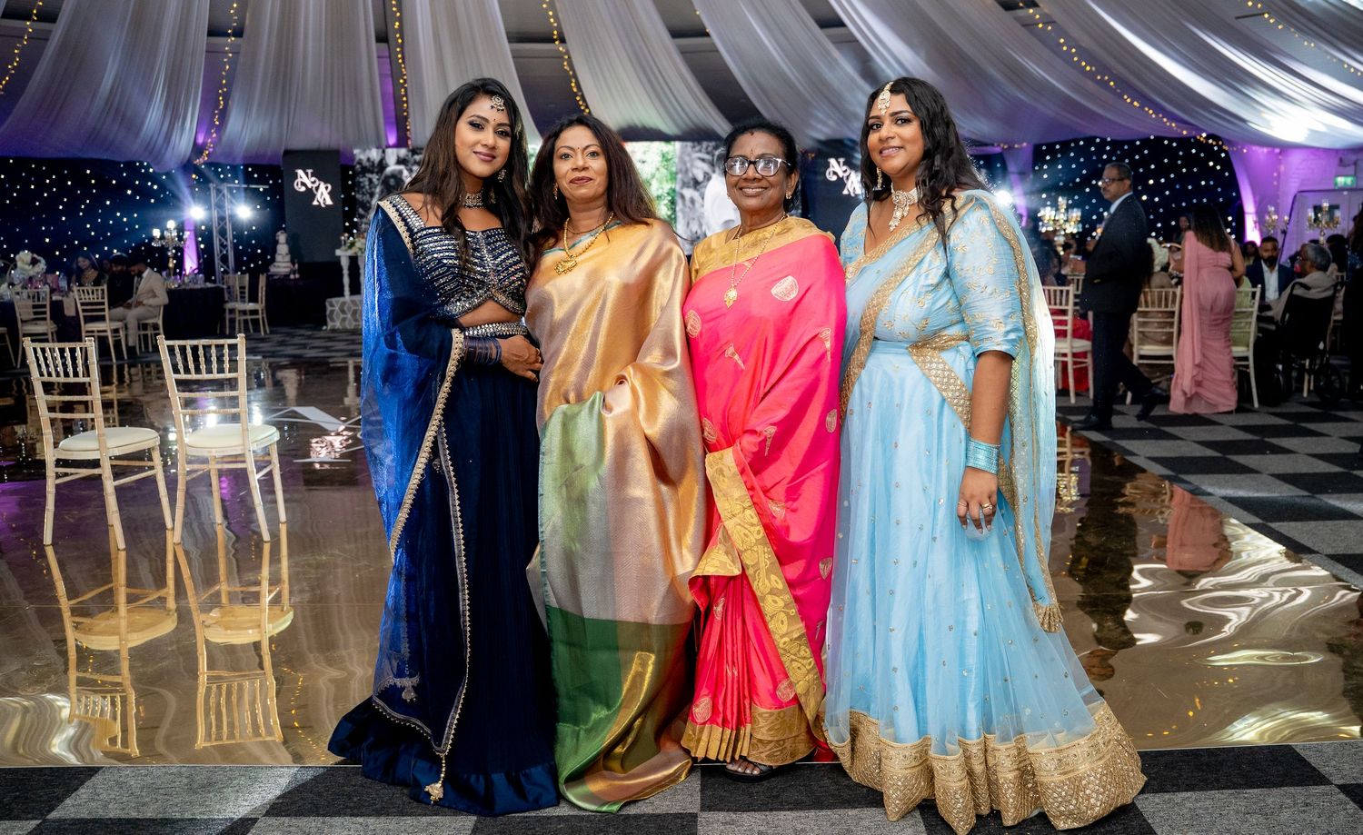Four women in elegant traditional and formal gowns pose together at an indoor evening celebration event.
