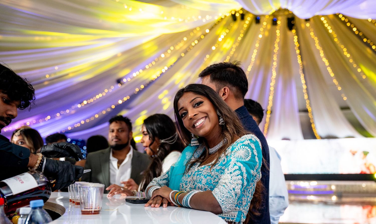 Smiling woman in turquoise outfit at wedding reception with decorative lighting and draped fabric ceiling.