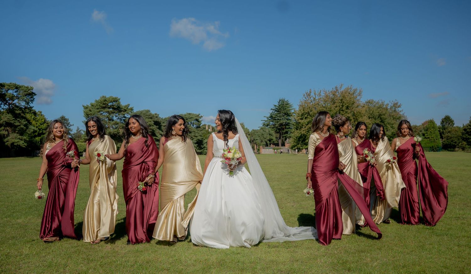 Wedding party walking on grass with bridesmaids in flowing burgundy and gold gowns beside bride.