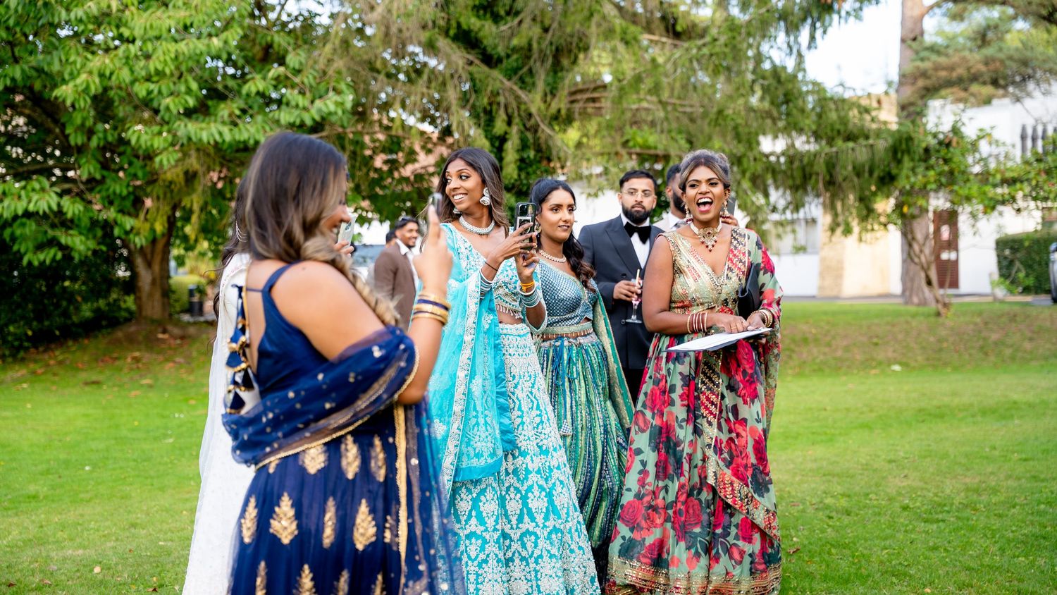 A group of women in vibrant Indian lehengas laugh and pose together on a lush green lawn.