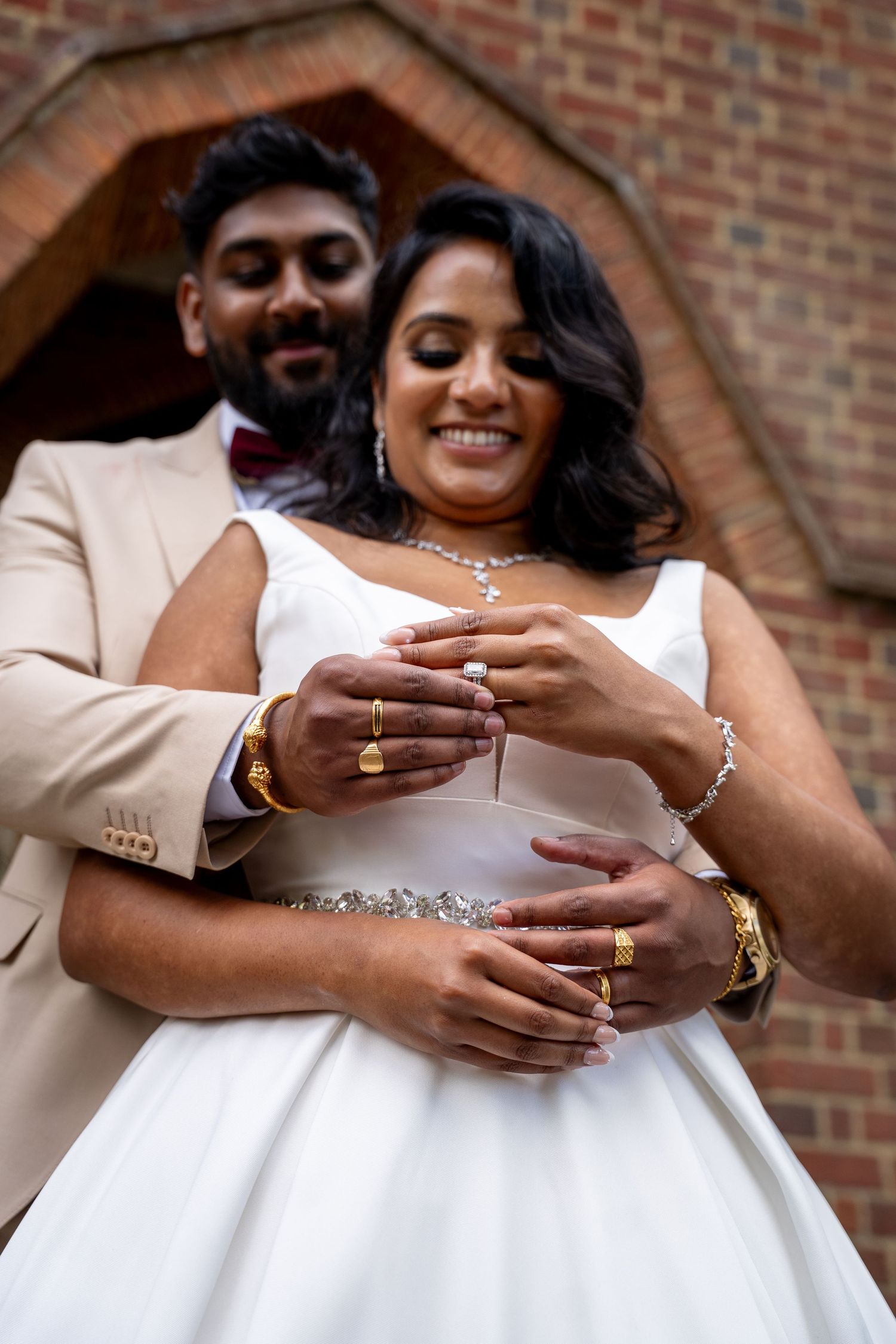Newlywed couple displaying wedding rings in front of brick archway, bride in white dress and groom in beige suit.