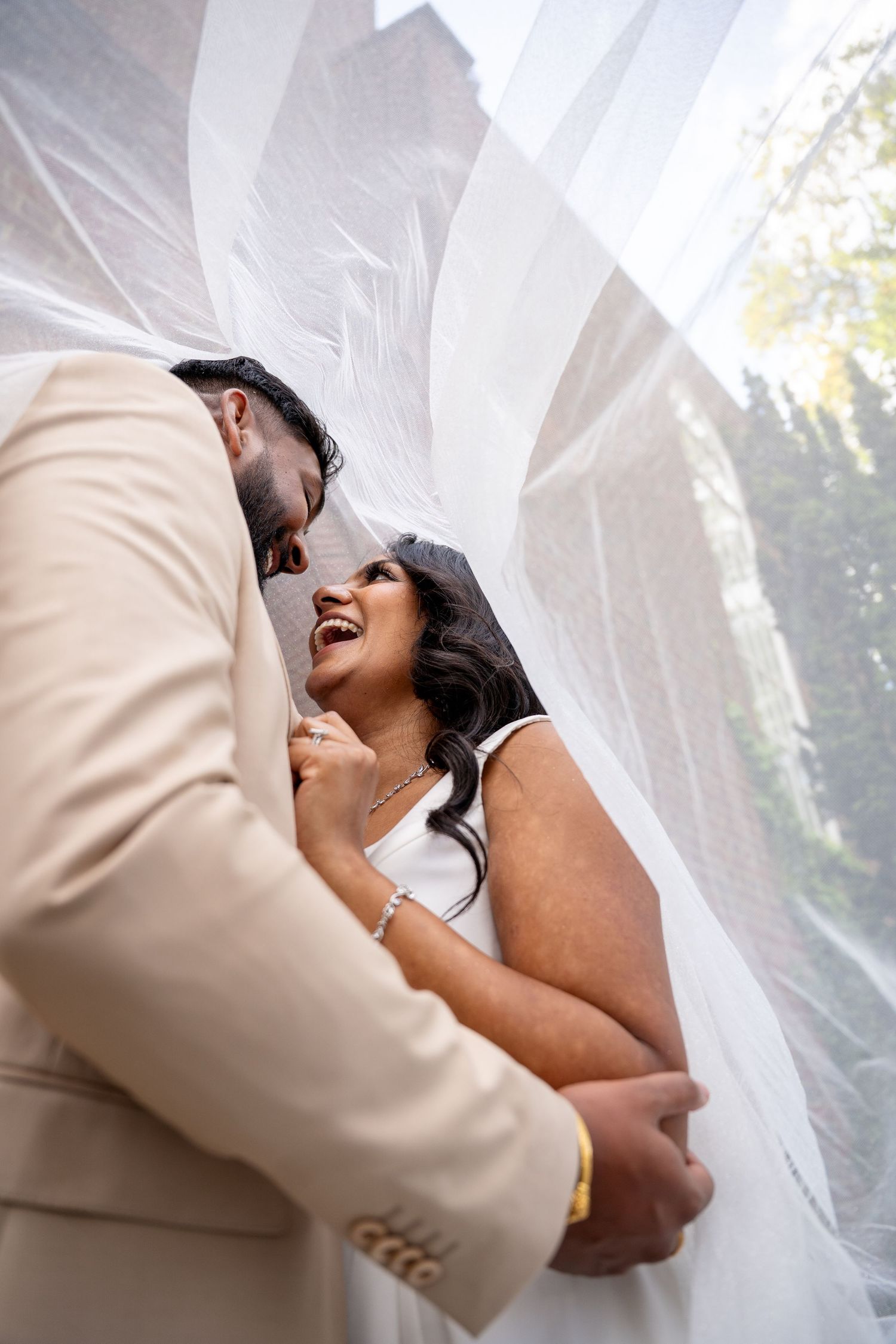 Bride laughing joyfully as groom whispers under flowing white veil outdoors.