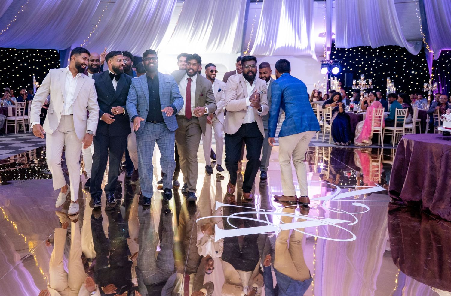 A group of men in formal attire gather and celebrate together on a mirrored Indian wedding dance floor.