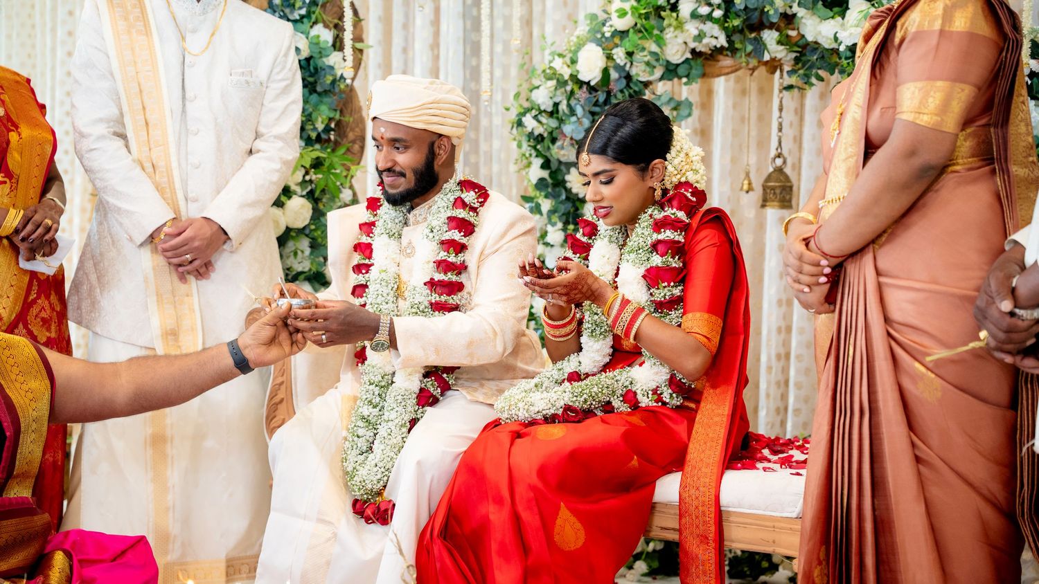 Guest presents item to couple during elaborate South Indian Hindu wedding ceremony on floral mandap.