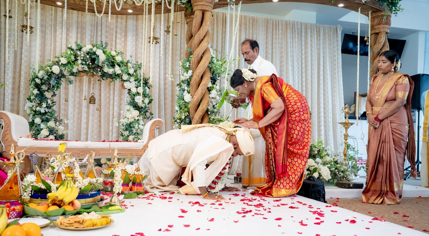 Groom bowing at feet of elders during traditional South Indian Hindu wedding ceremony with floral decorations.