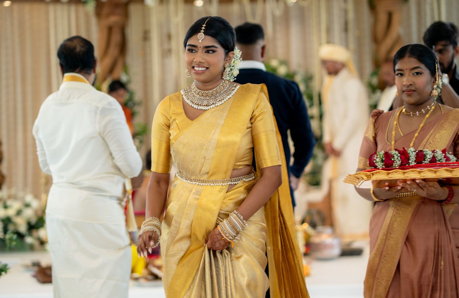 Bride in yellow silk saree with diamond necklace walks through decorated South Indian wedding venue.