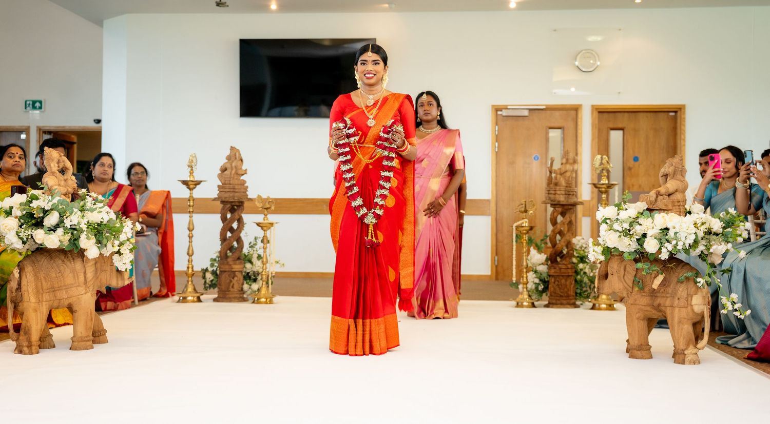 South Indian bride in red silk saree processing through wedding venue flanked by brass oil lamps.