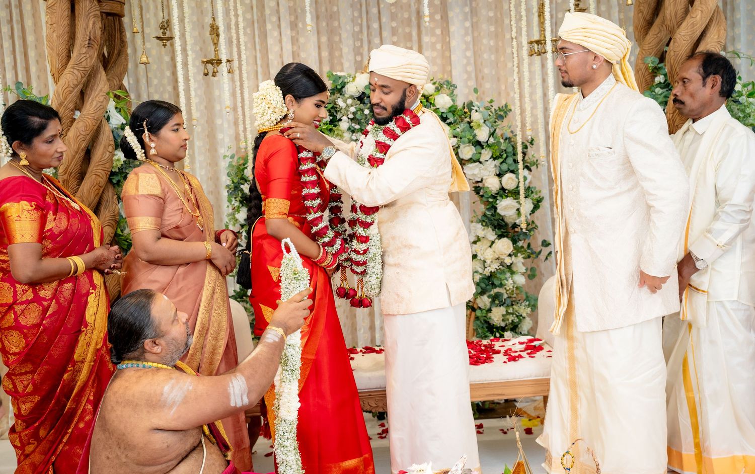 Traditional South Indian wedding garland exchange ceremony with priest, bride in red saree and groom in white.