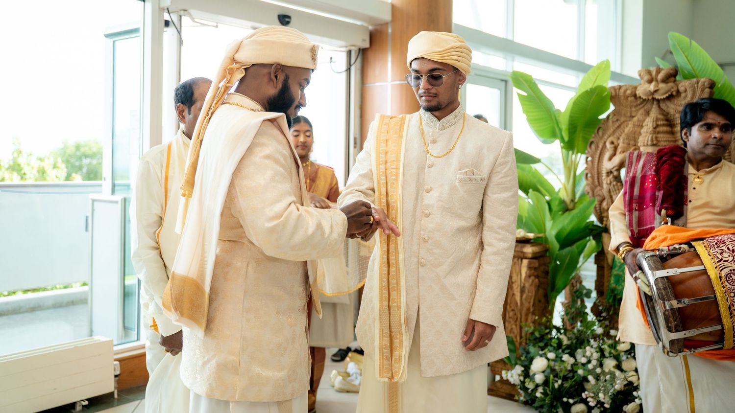 Two men in cream sherwanis and golden turbans sharing a warm handshake during a traditional Hindu wedding ceremony.