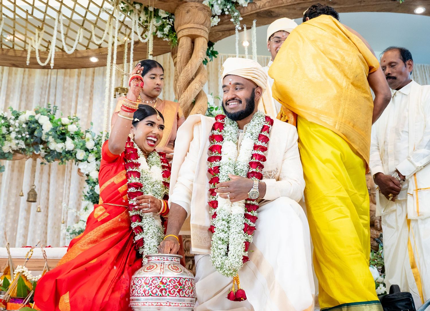 Laughing bride and groom exchange garlands during a joyful traditional South Indian Hindu wedding ceremony.