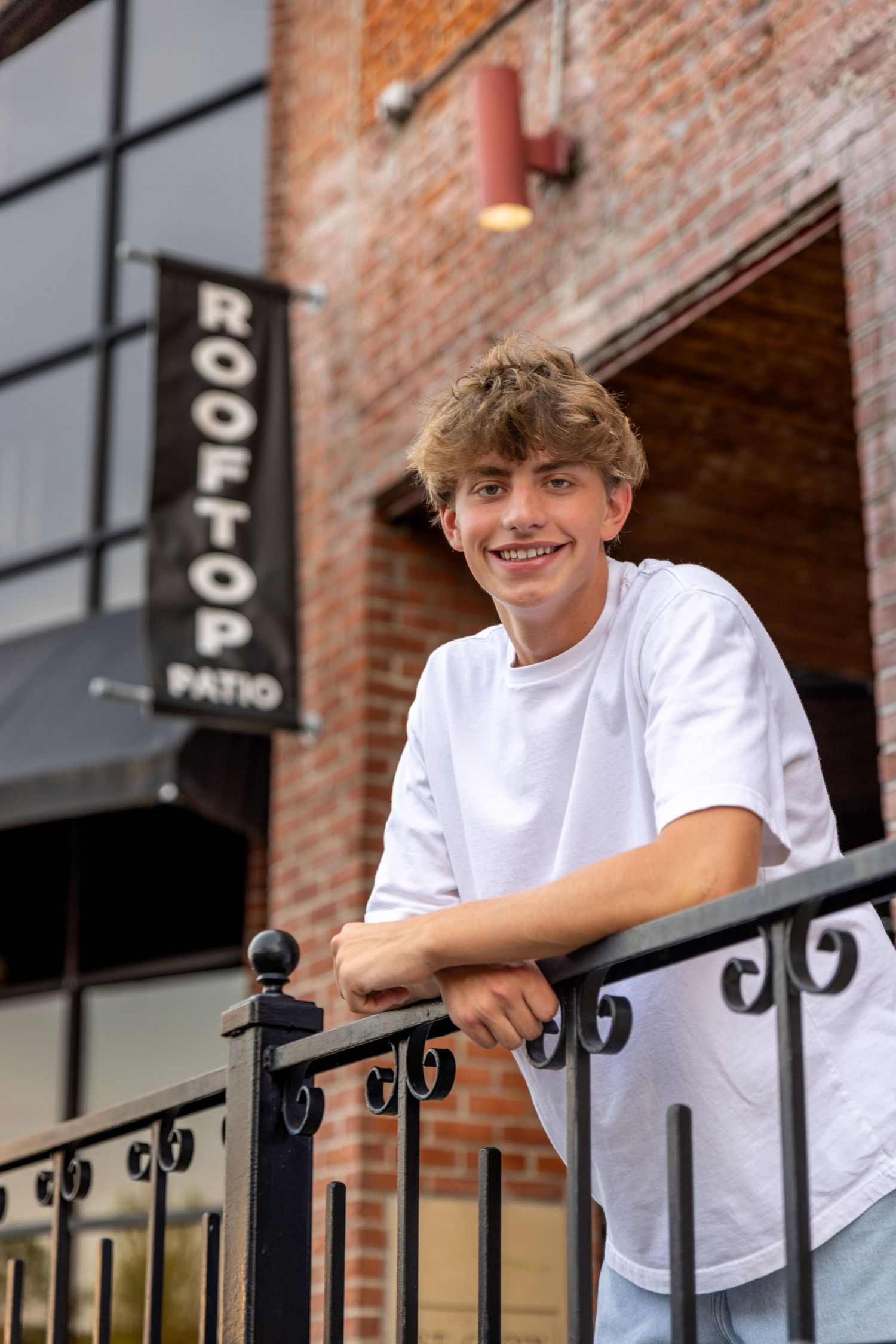 Smiling teenage boy leaning on black iron railing outside a brick building with a rooftop patio sign.