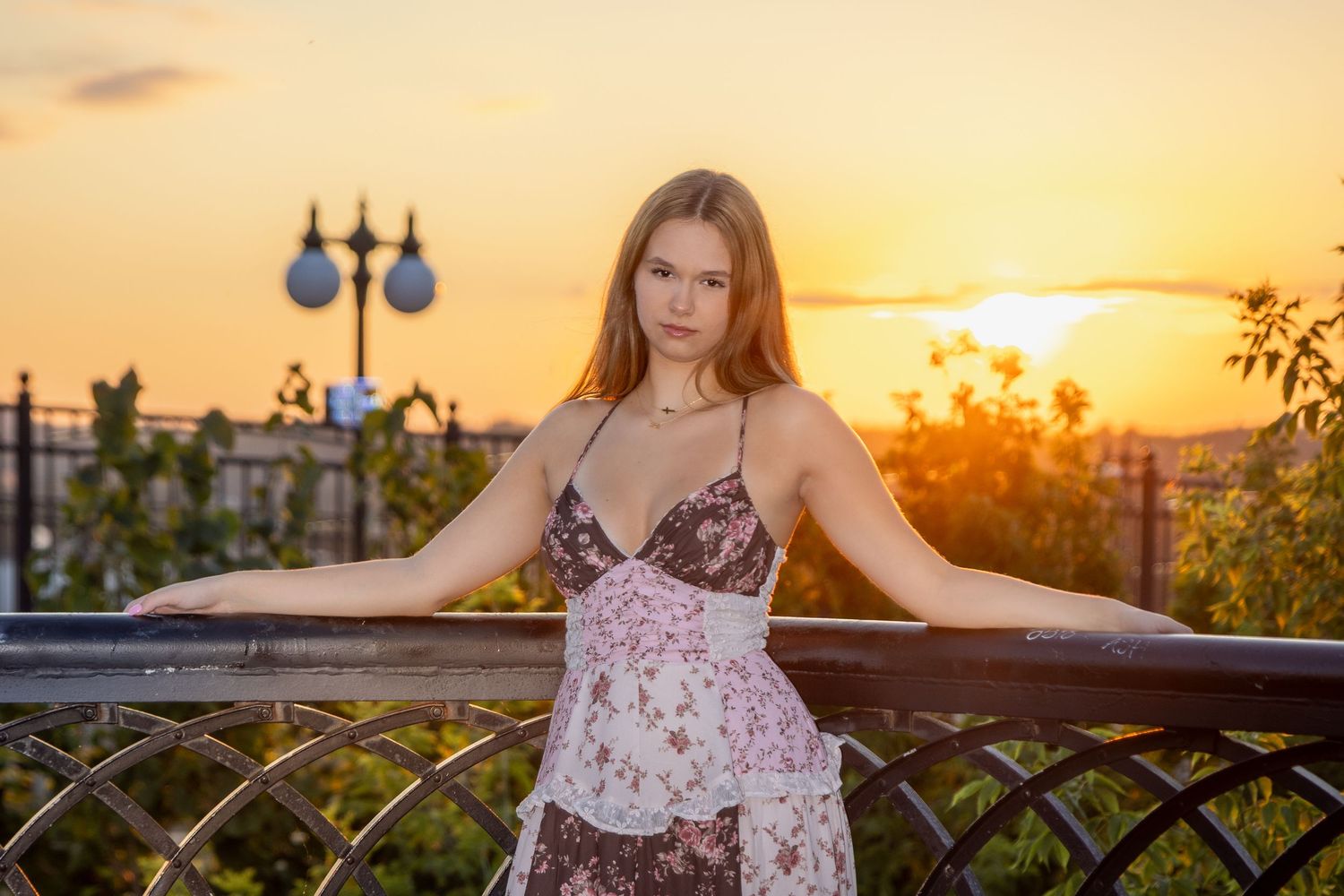 Young woman in floral dress poses by iron railing at golden sunset, city lights glowing in warm evening sky.